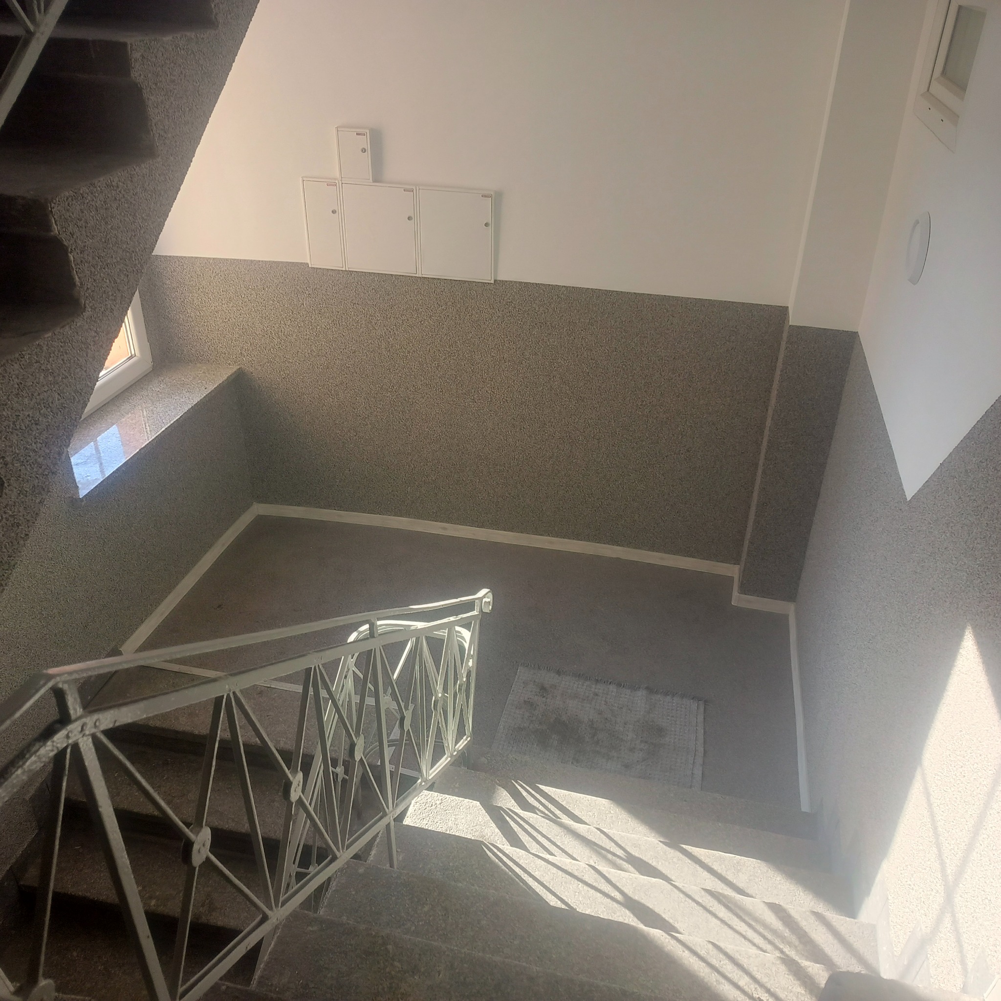 Klatka schodowa z szarymi ścianami o ziarnistej fakturze, białe szafki na ścianie i metalowa balustrada. Widok z góry na schody oświetlone naturalnym światłem.