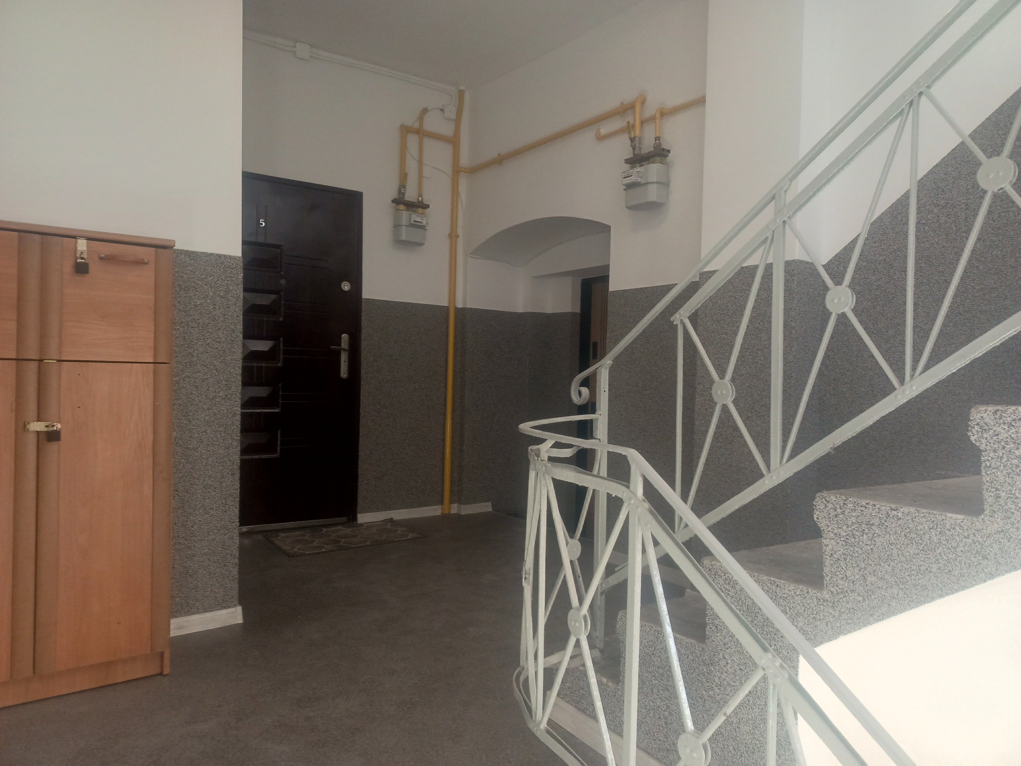 Klatka schodowa w bloku z drewnianą szafką, metalowymi rurami gazowymi i schodami z szarą, kamienną fakturą. Białe ściany i drzwi wejściowe z numerem 5.