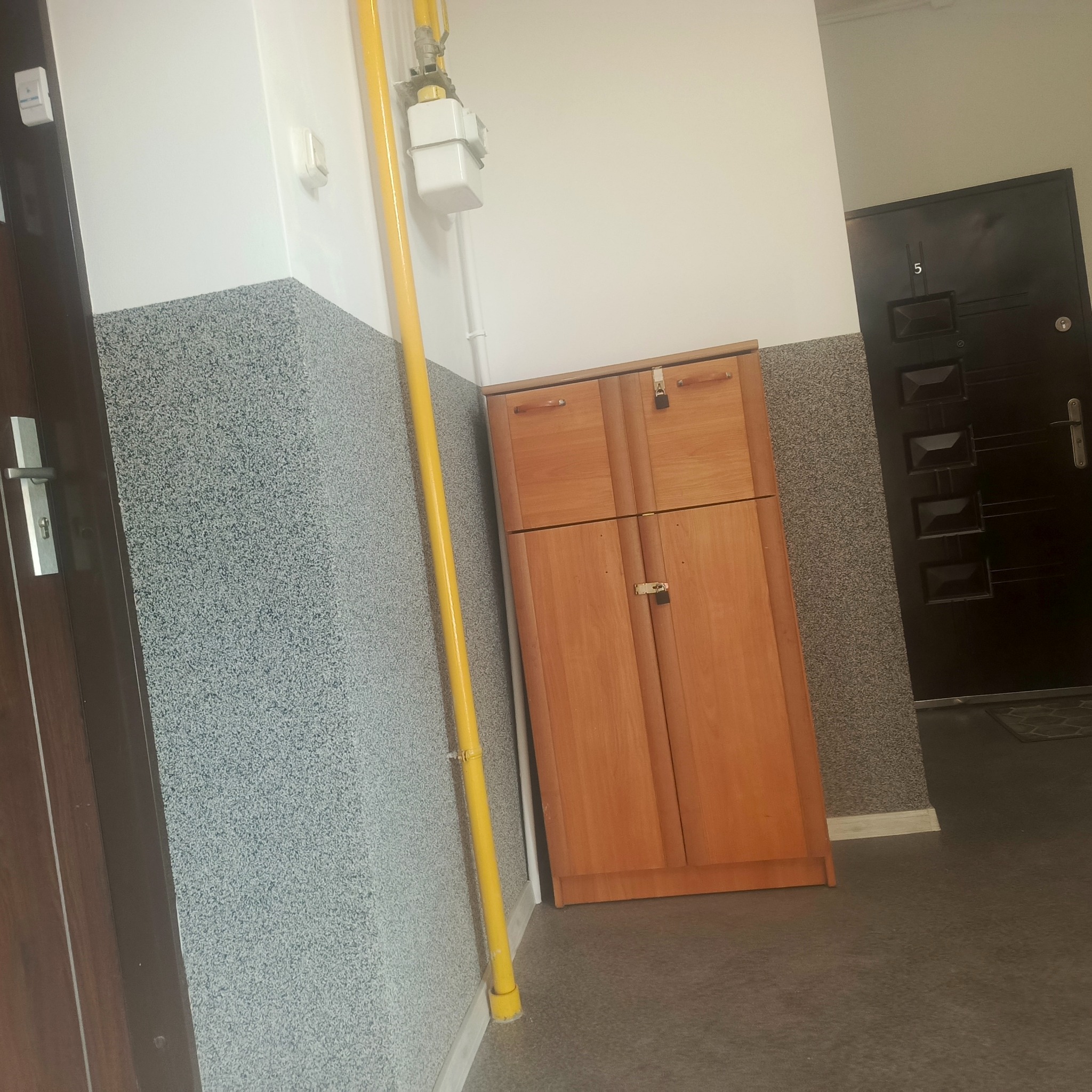 Korytarz z szafką na buty, żółtą rurą gazową i drzwiami wejściowymi z numerem 5. Ściany wykończone w dolnej części szarym, wzorzystym tynkiem.