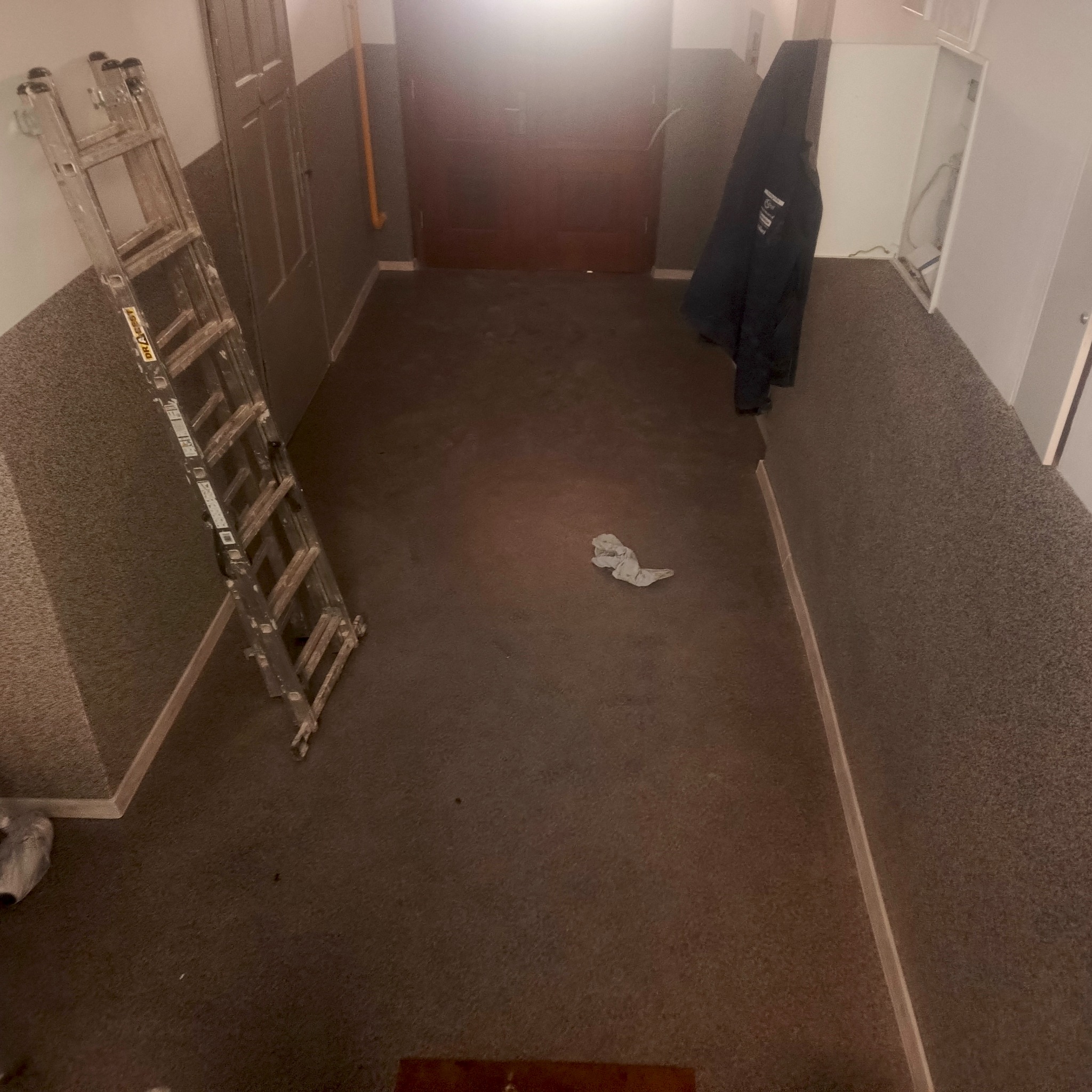 Korytarz w trakcie remontu: drabina oparta o ścianę, ubranie wiszące na wieszaku, ślady po pracy na podłodze. Przygotowania do malowania lub wykończenia.