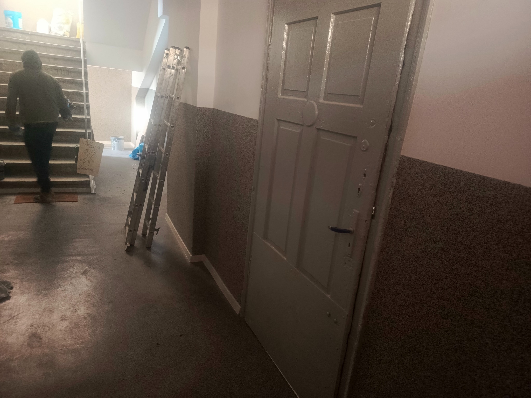 Klatka schodowa w trakcie malowania: drabina oparta o ścianę, pomalowane drzwi, osoba wchodząca po schodach. Widoczne ślady farby na podłodze.
