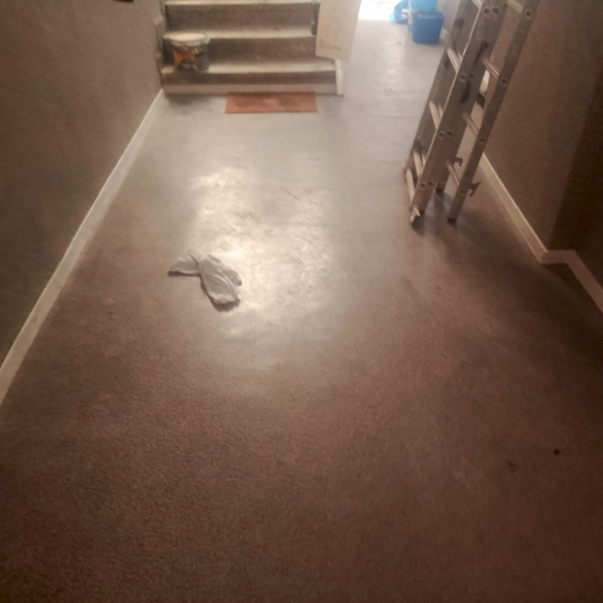 Wnętrze w trakcie prac wykończeniowych: świeżo wylana, brązowa posadzka, drabina oparta o ścianę, rękawice robocze leżące na podłodze, schody w tle.