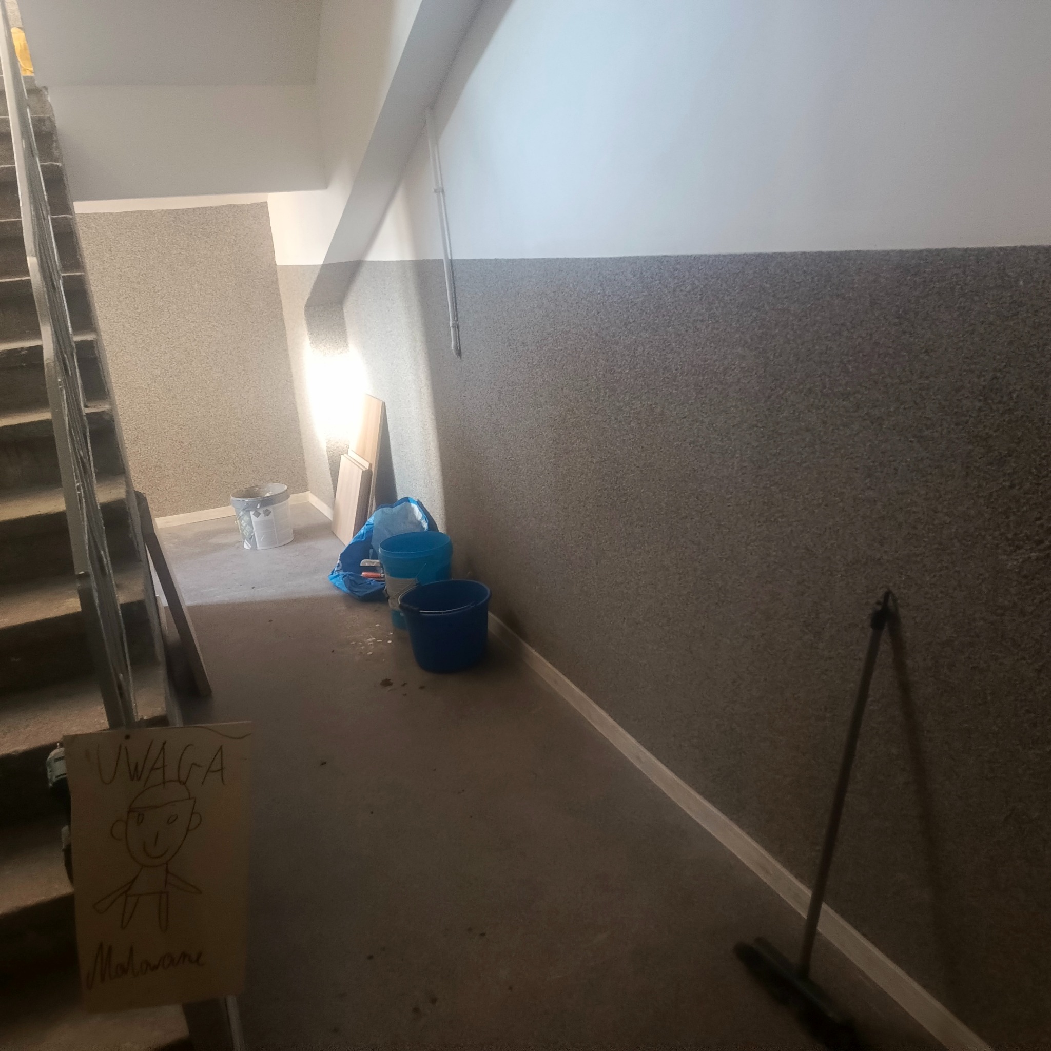 Klatka schodowa w trakcie malowania: widoczne wiadra z farbą, drabina, kartka z napisem 'Uwaga Malowane' i rysunkiem. Ściany pomalowane do połowy na szaro.