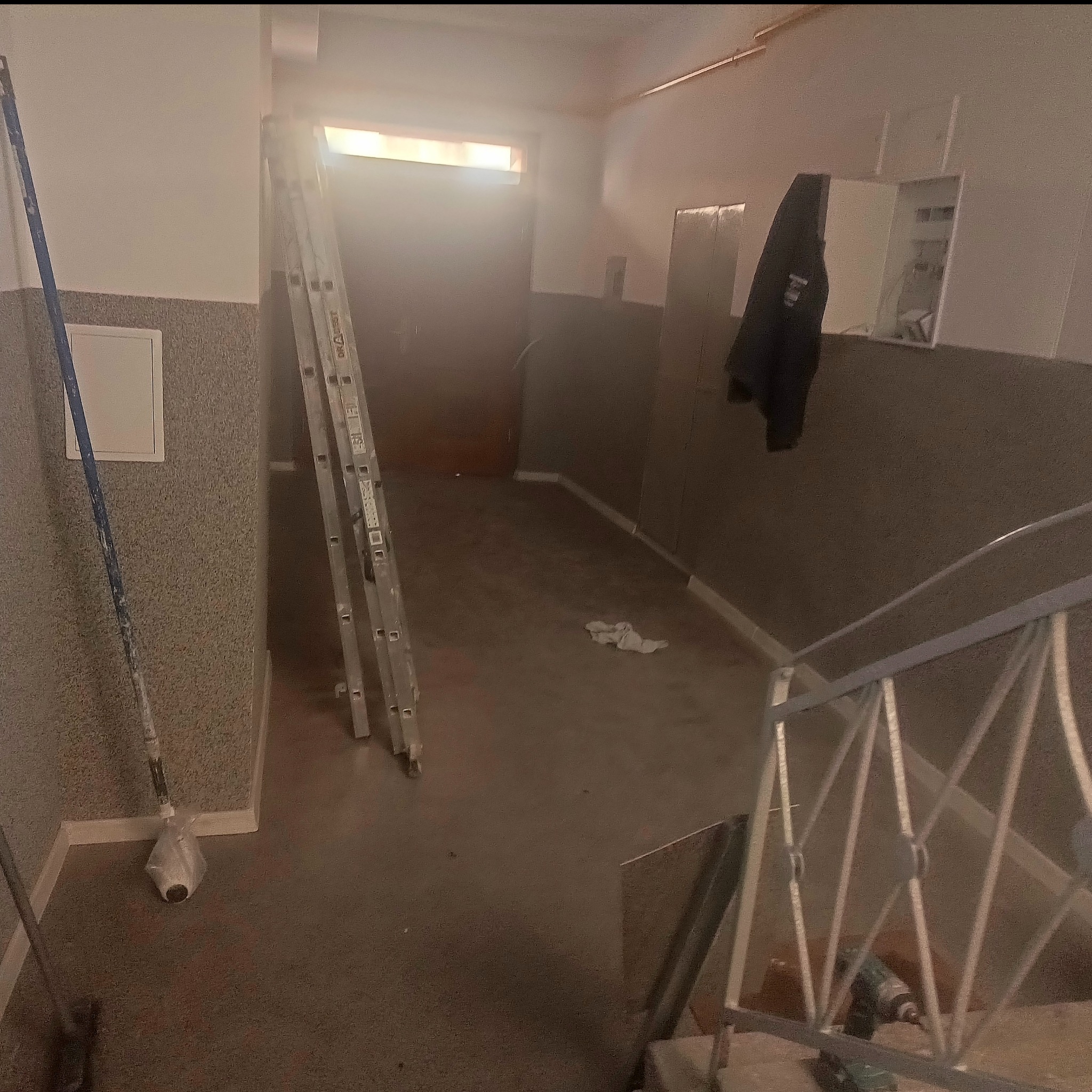 Klatka schodowa w trakcie prac wykończeniowych: drabina oparta o ścianę, wałek malarski, w tle drzwi i schody z metalową barierką. Widoczne ślady prac remontowych.