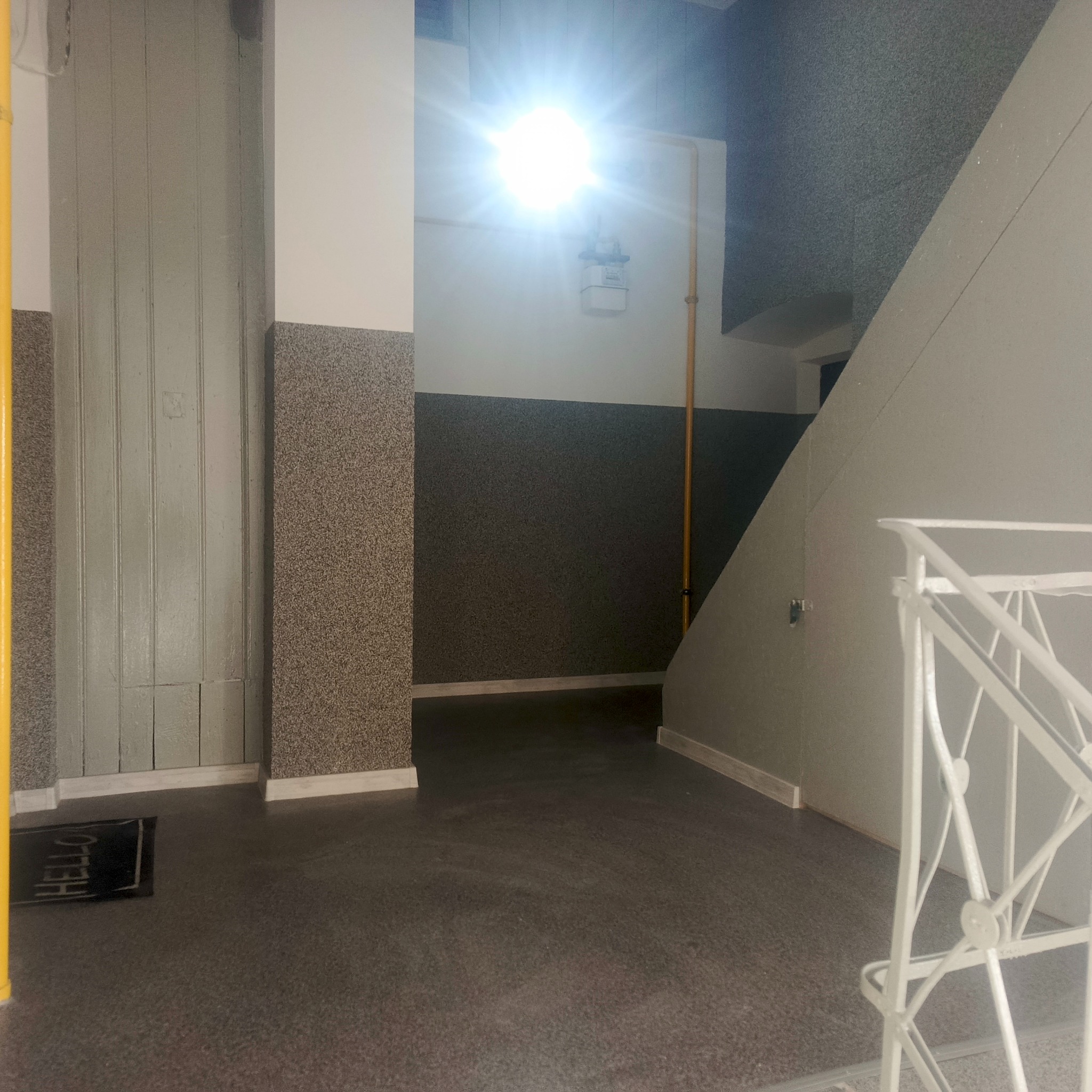 Klatka schodowa w bloku z szarą podłogą, ścianami w dwóch odcieniach szarości i białą balustradą. Widoczny fragment drzwi i rura gazowa.