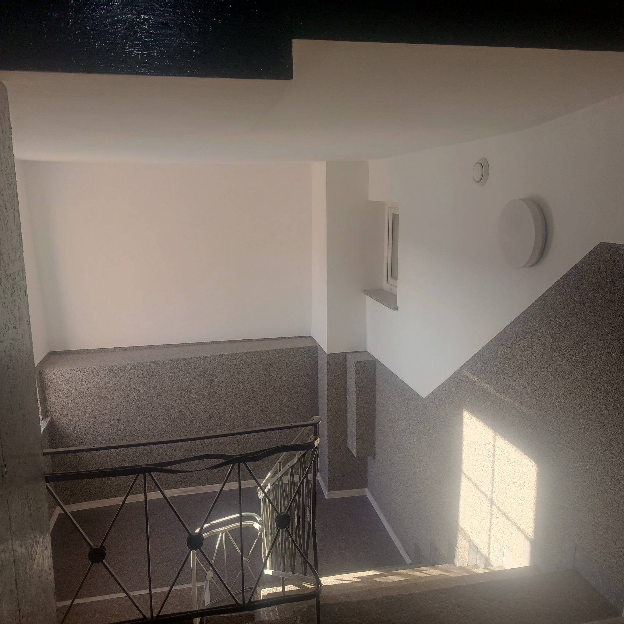 Klatka schodowa po remoncie: białe ściany, szare wykończenie schodów i podestów, czarna balustrada. Widok z góry, światło słoneczne wpadające przez okno.