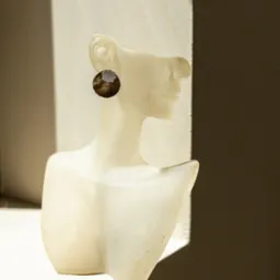 Kolczyk z brązowo-srebrnym wzorem na białym manekinie w oknie, oświetlony naturalnym światłem, z widocznym cieniem na ścianie.