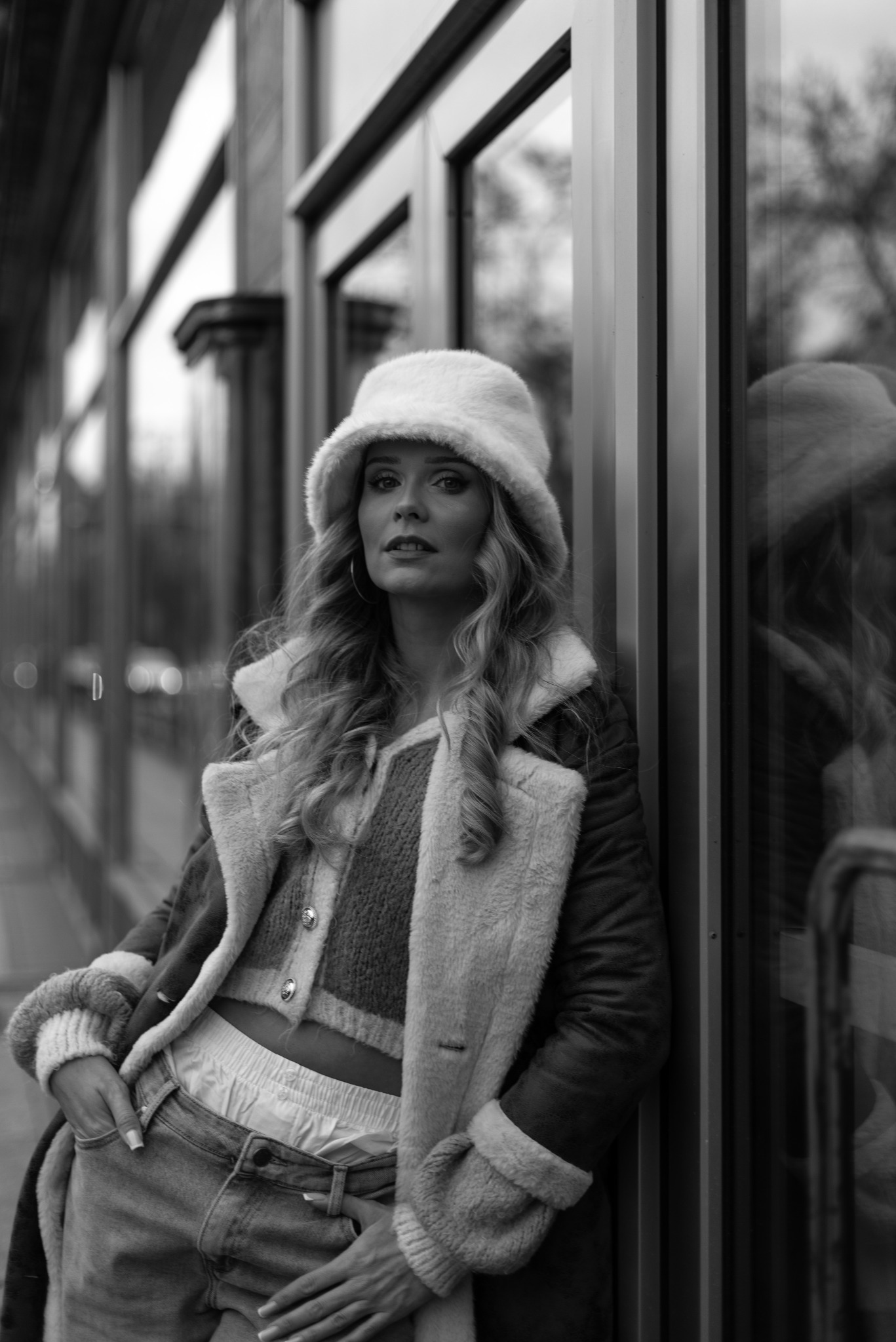 Czarno-biała sesja portretowa kobiety w futrzanym kapeluszu i płaszczu z kożuszkiem, oparta o szklaną ścianę budynku. Widoczne odbicie w szybie.