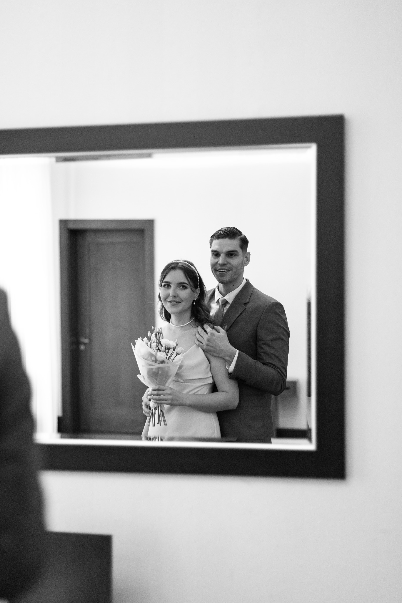 Czarno-białe odbicie w lustrze: uśmiechnięta para w strojach ślubnych, kobieta trzyma bukiet. Mężczyzna obejmuje kobietę. Widoczne drzwi w tle.