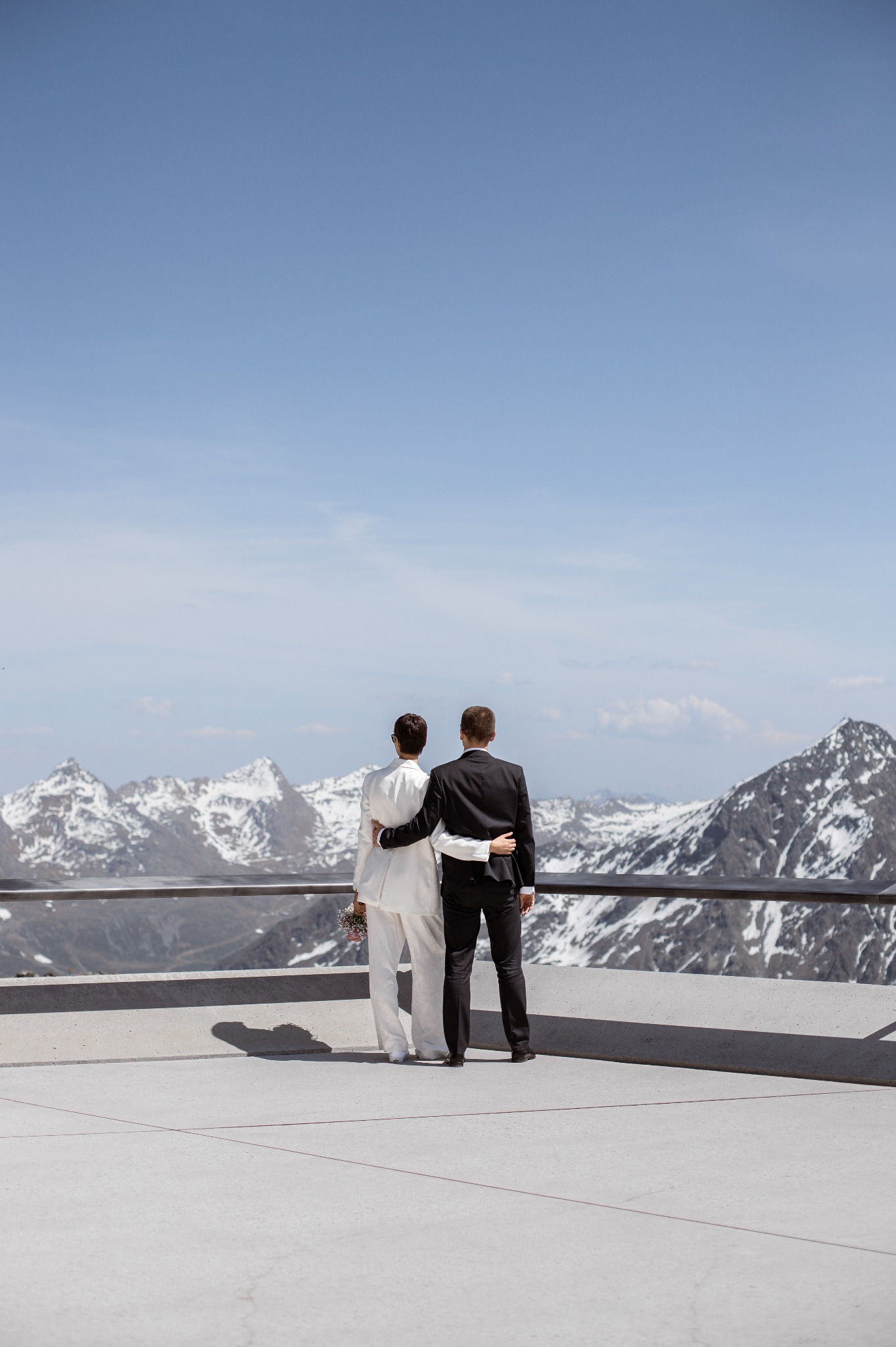 Para młoda, tyłem, obejmuje się na tle górskich szczytów. Ona w białym garniturze, on w czarnym. Minimalistyczny kadr, jasne tło, nowoczesna architektura.