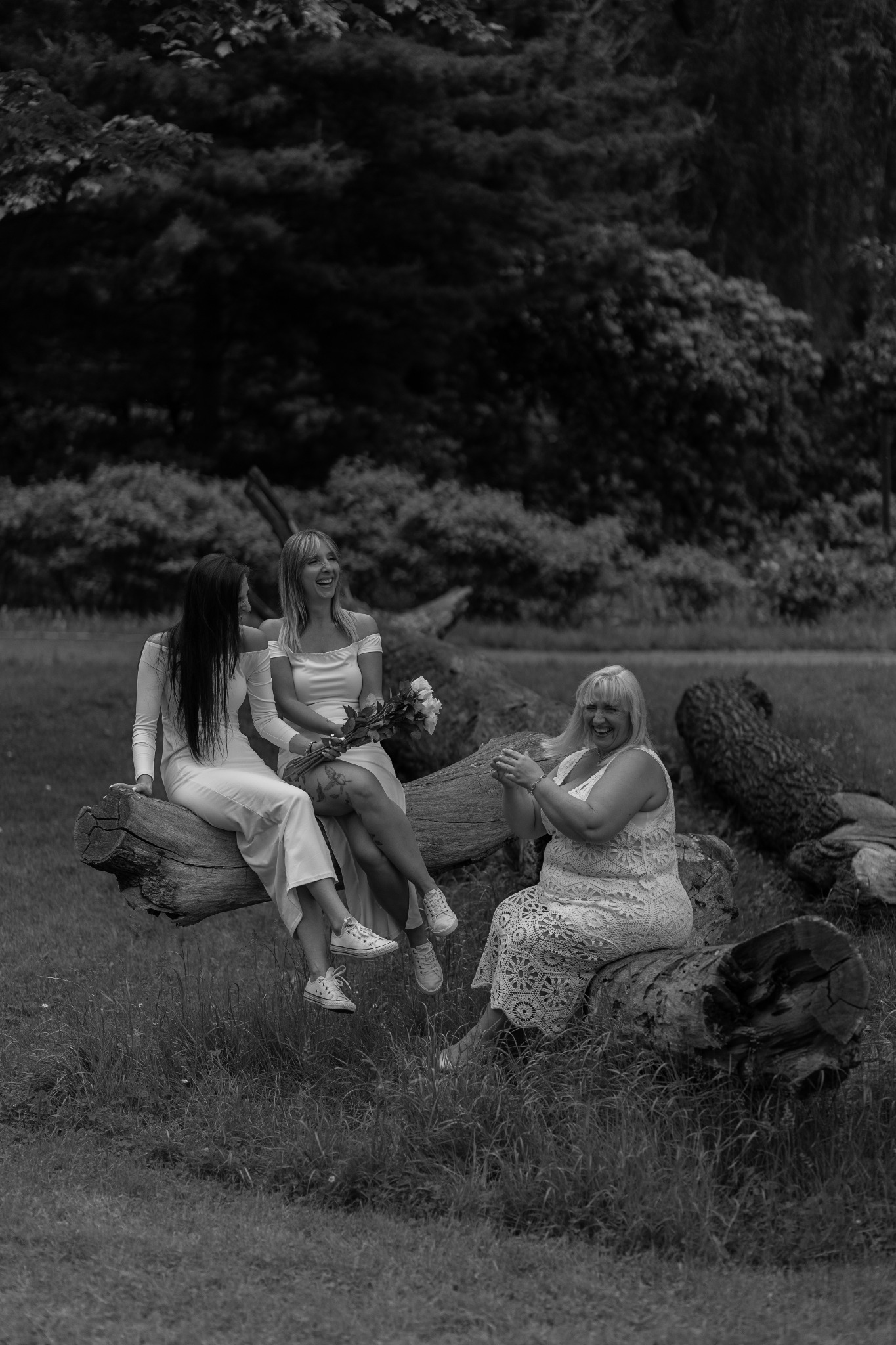 Trzy kobiety w jasnych sukienkach siedzą na powalonych pniach drzew w plenerze. Jedna z kobiet trzyma bukiet kwiatów. Zdjęcie czarno-białe, o naturalnym oświetleniu.