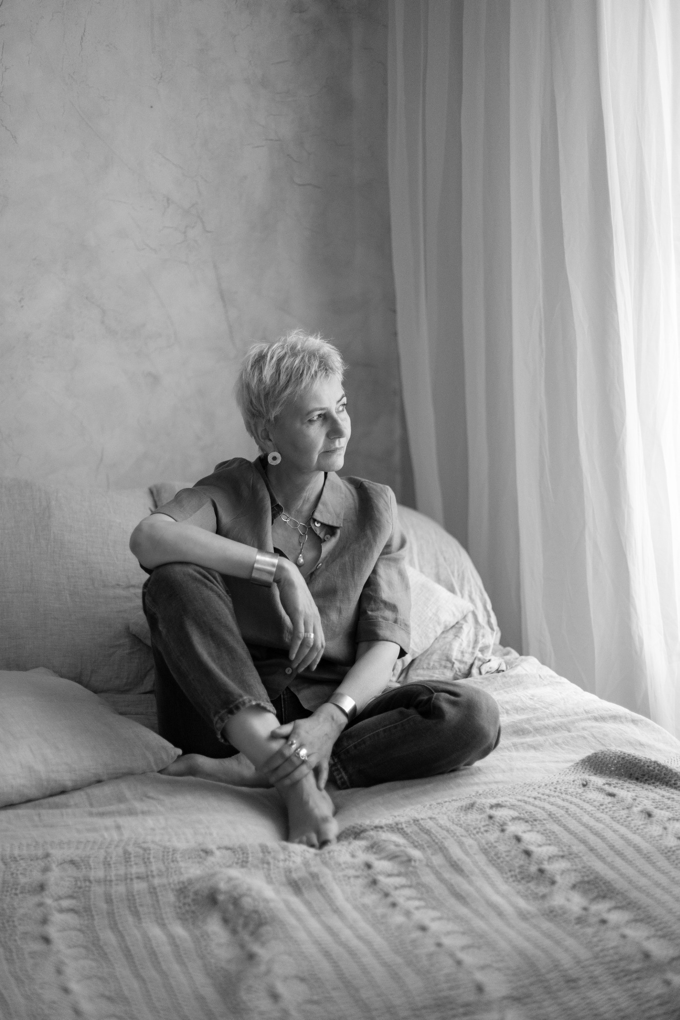 Portret kobiety z krótkimi włosami, siedzącej na łóżku przy oknie, w czarno-białej tonacji. Ubrana w koszulę i dżinsy, z biżuterią. Spokojny wyraz twarzy.
