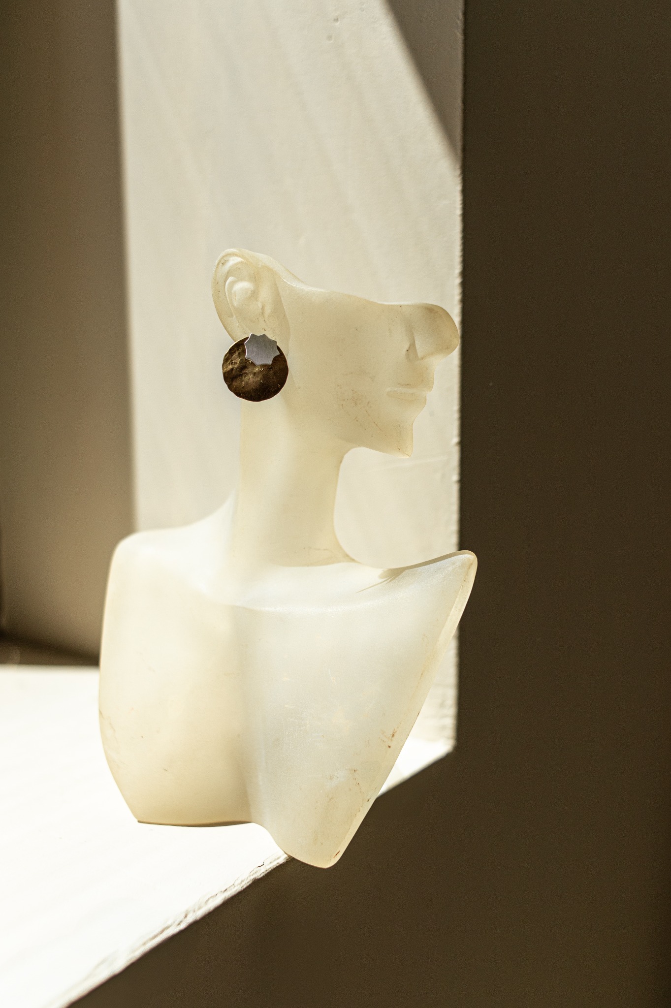 Kolczyk z brązowo-srebrnym wzorem na białym manekinie w oknie, oświetlony naturalnym światłem, z widocznym cieniem na ścianie.