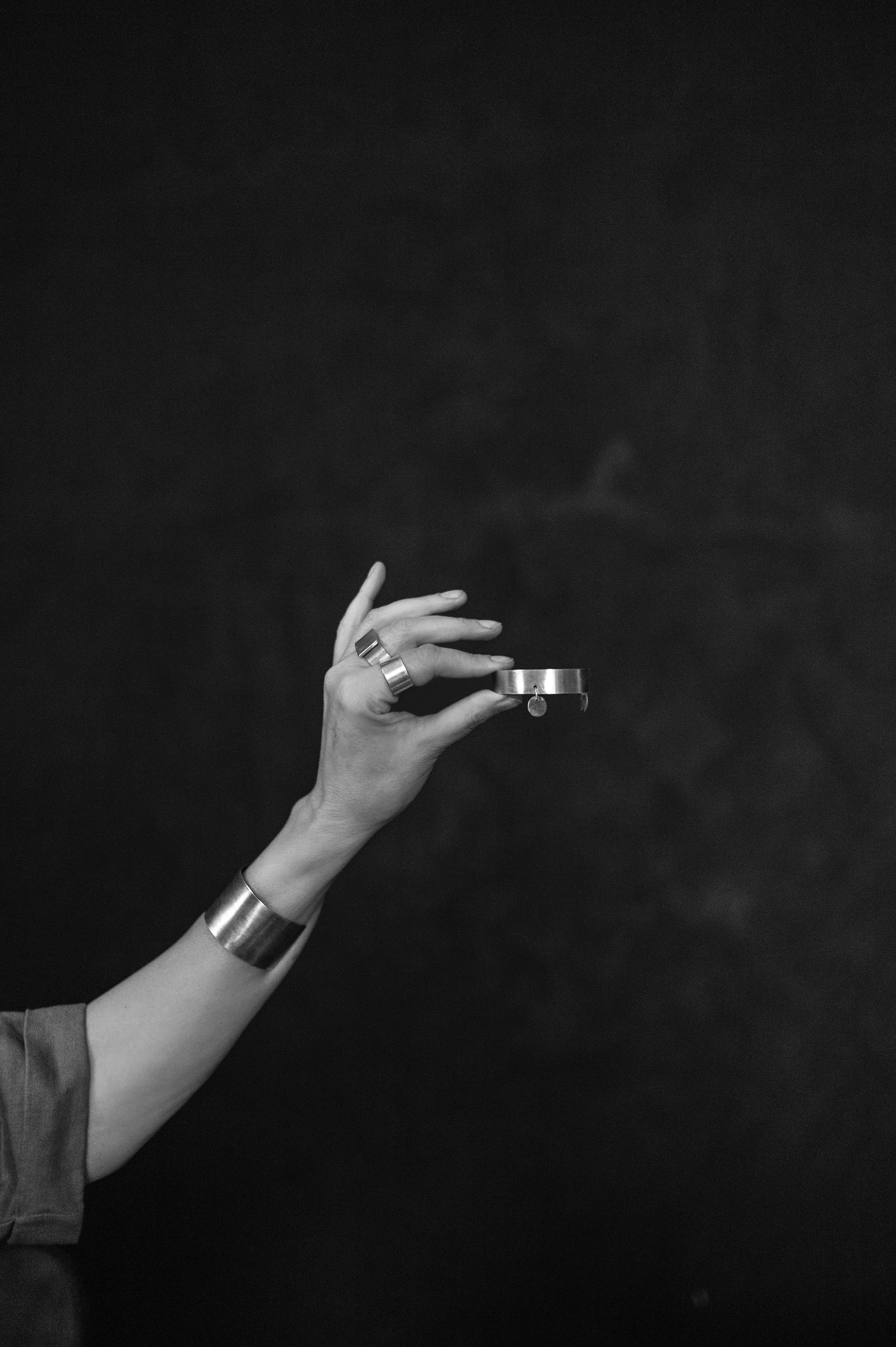 Czarno-białe zdjęcie dłoni z biżuterią (pierścionki, bransoletka na nadgarstku i w dłoni) na ciemnym tle. Minimalistyczny styl, ostre światło.