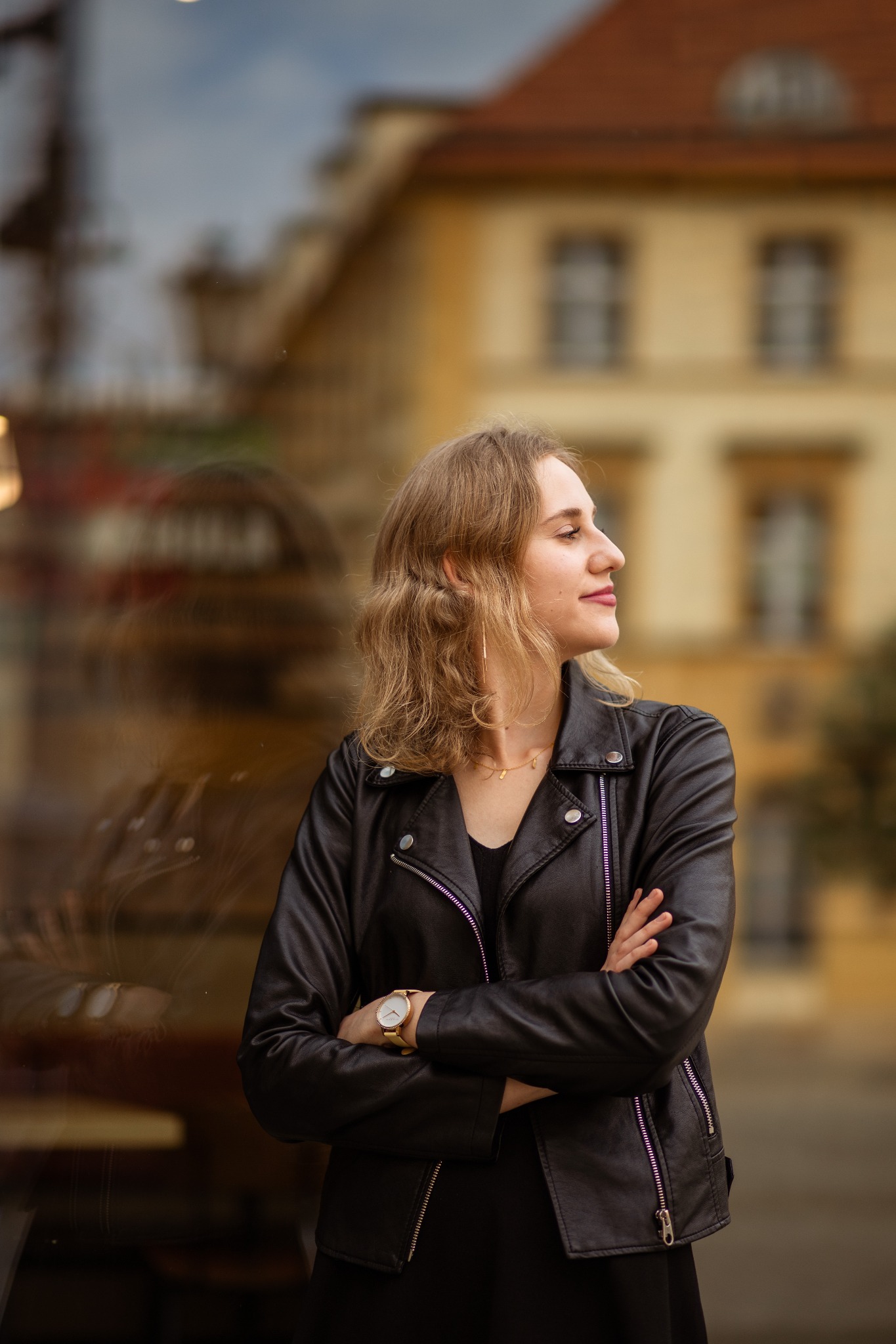 Portret kobiety w czarnej skórzanej kurtce, z założonymi rękami, stojącej na tle rozmytego budynku z cegły. Złoty zegarek na nadgarstku.