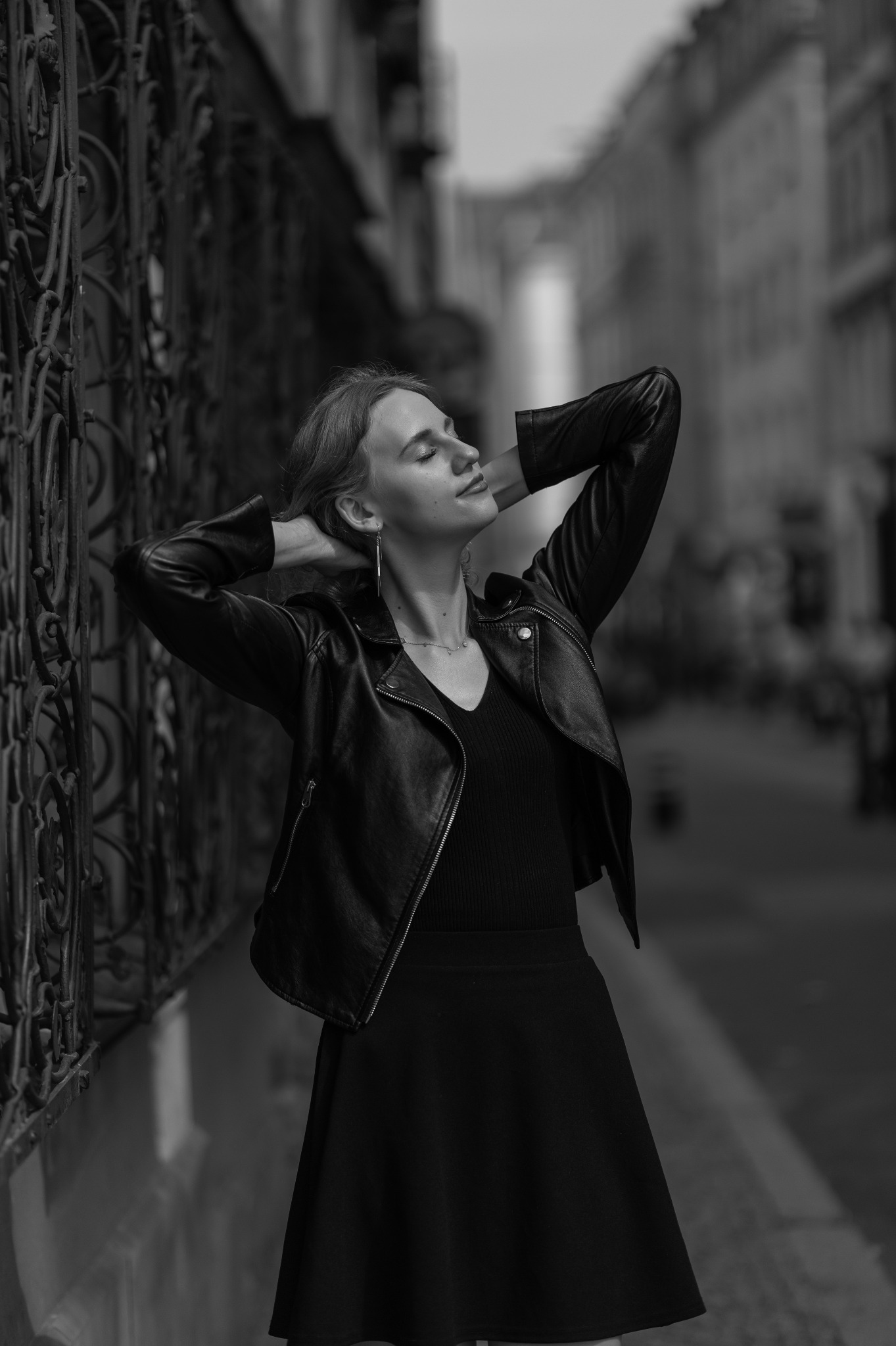 Stylowe, czarno-białe zdjęcie kobiety w skórzanej kurtce, opierającej się o ozdobną, metalową balustradę, z zamkniętymi oczami i uniesionymi rękami na tle ulicy.