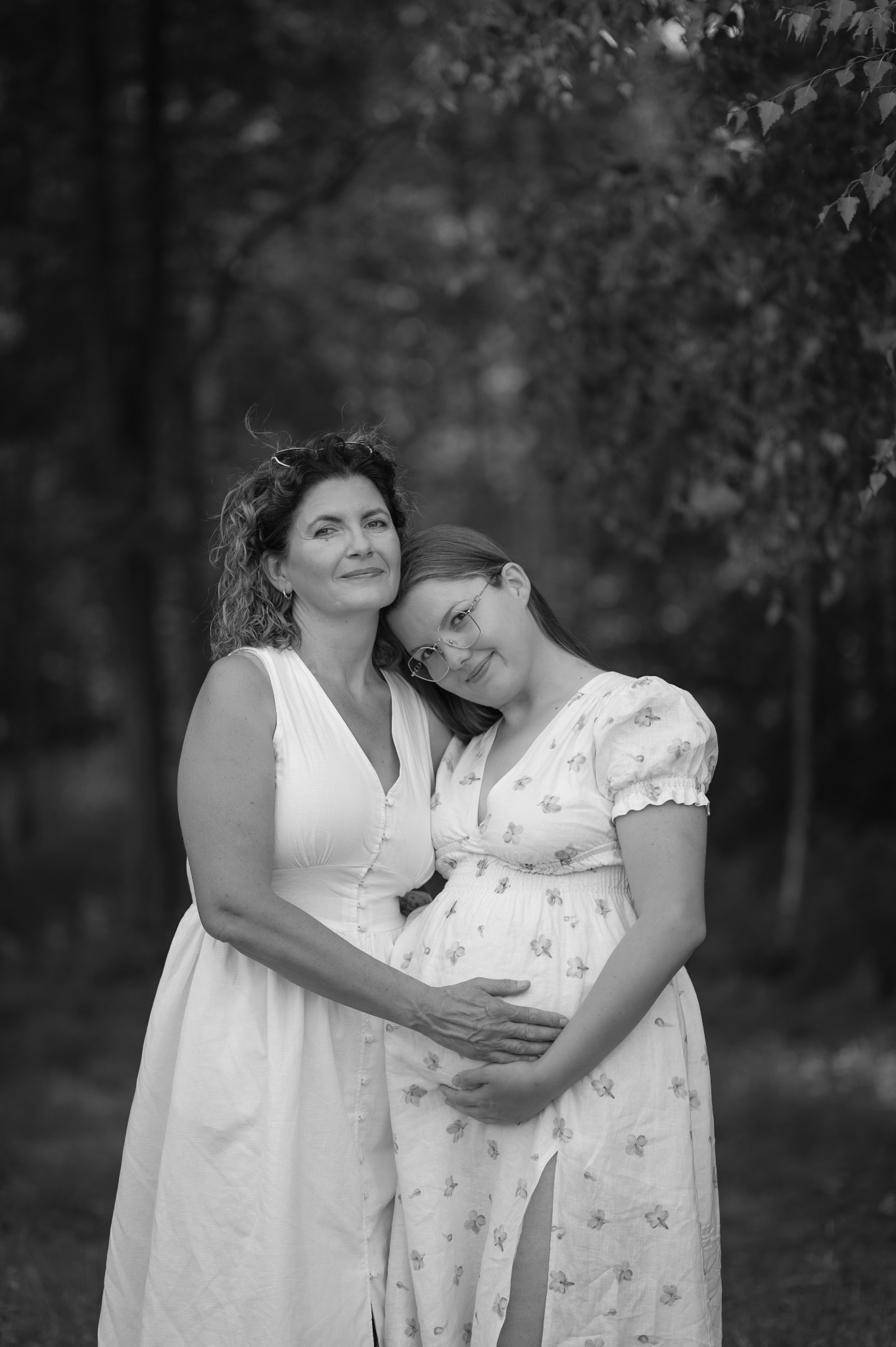 Czarno-białe zdjęcie matki i córki w ciąży, tulących się na tle zieleni we Wrocławiu. Córka w kwiecistej sukience, matka w białej. Delikatne światło podkreśla emocje.