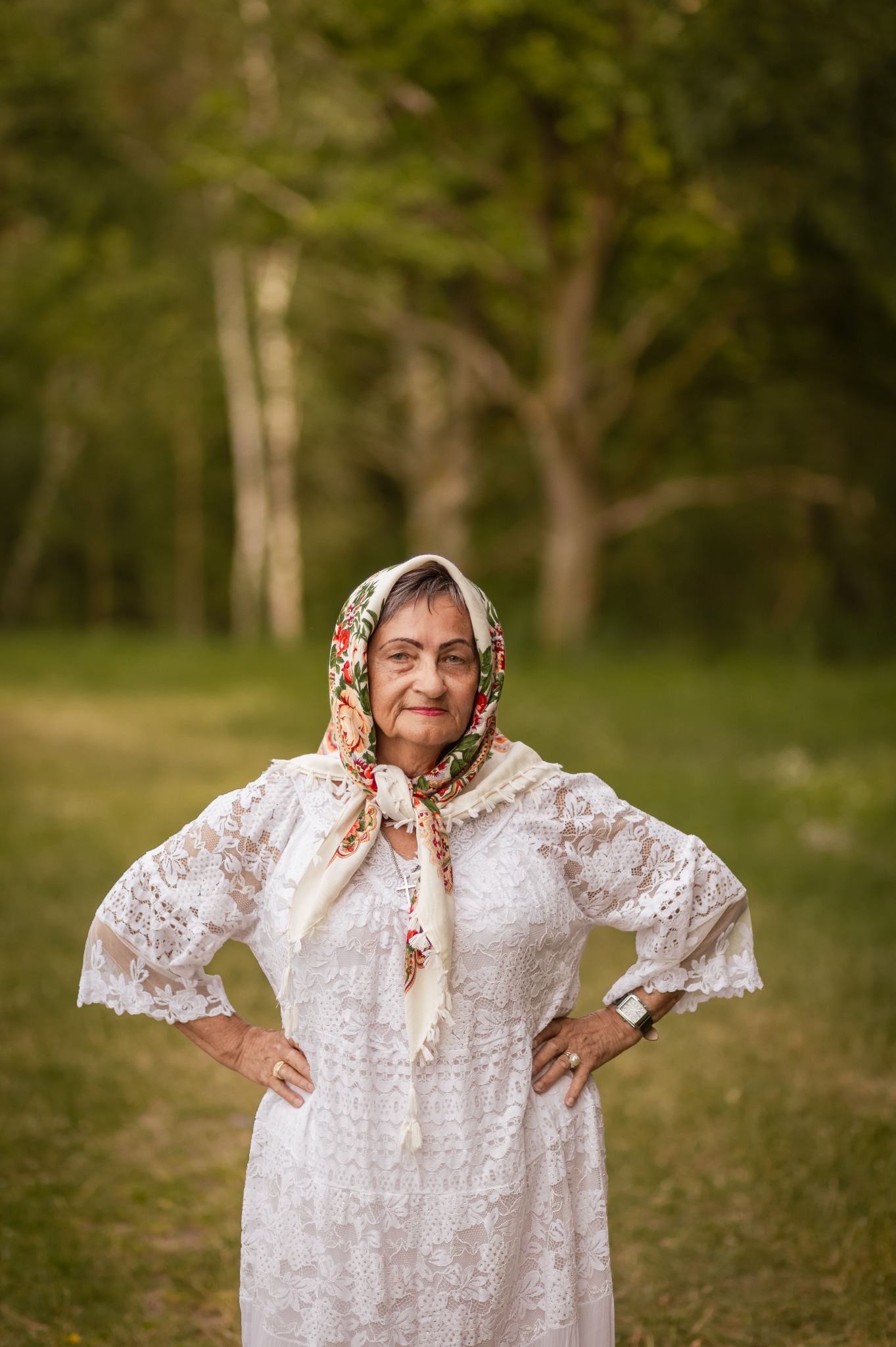 Portret starszej kobiety w białej, koronkowej sukience i kwiecistej chuście na głowie, pozującej na tle zieleni. Widoczne detale: krzyżyk na szyi, zegarek na ręce.