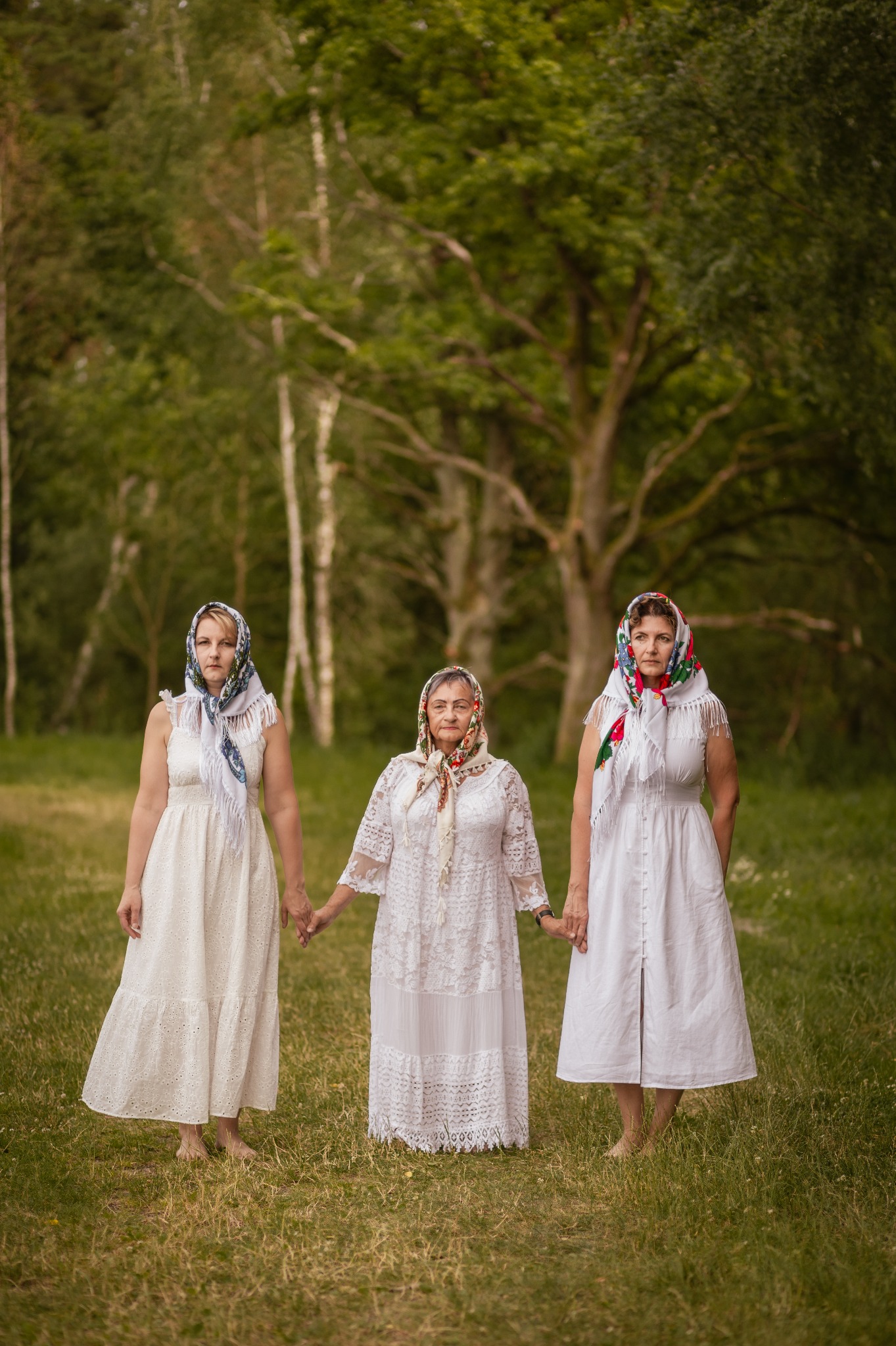 Trzy kobiety w białych sukienkach i chustach na głowach, stojące na trawie na tle drzew. Kobiety trzymają się za ręce. Zdjęcie w plenerze, w naturalnym oświetleniu.