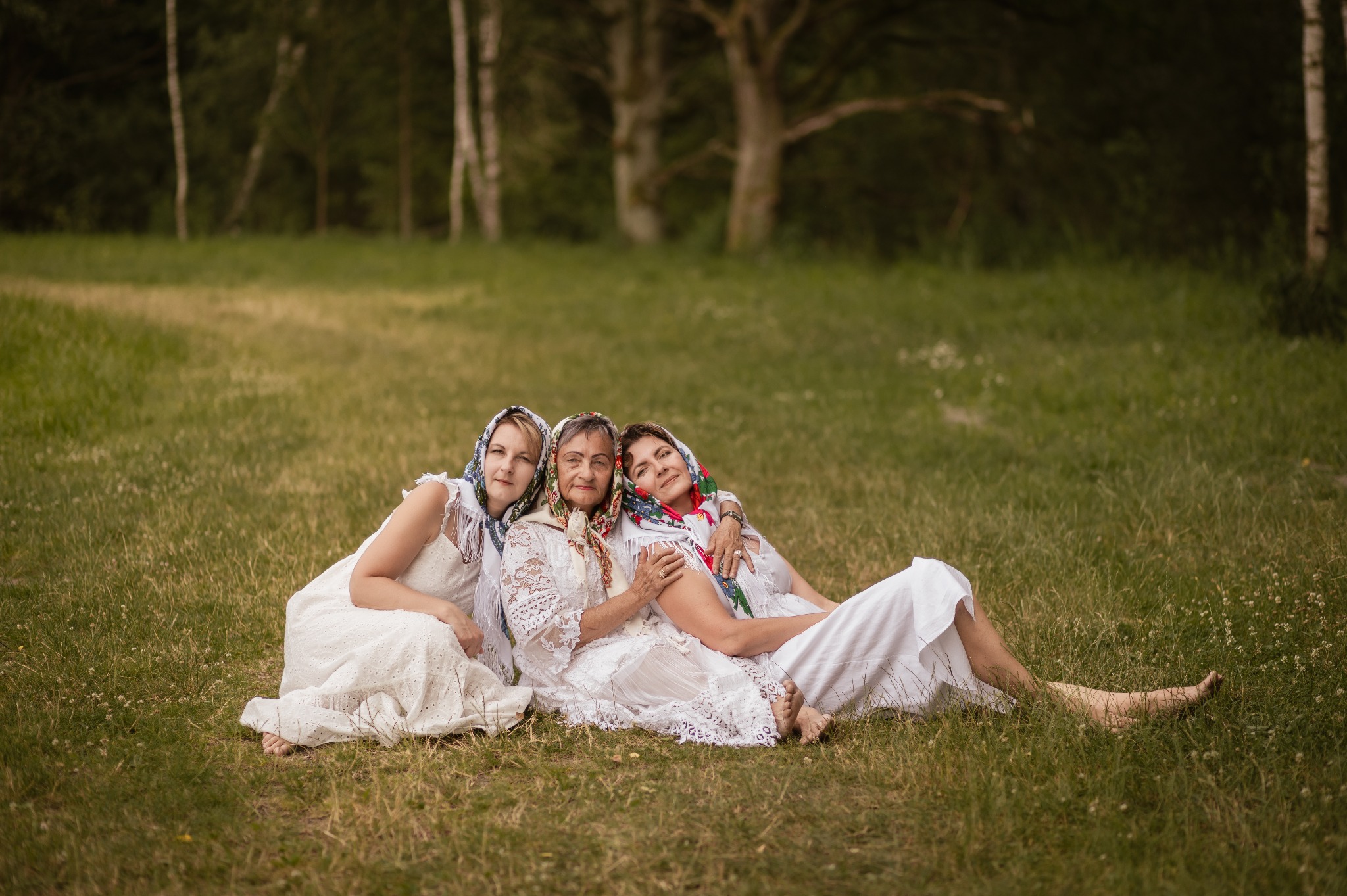 Trzy kobiety w białych sukienkach i chustach na głowach siedzą na trawie w plenerze. Uśmiechnięte twarze, naturalne światło, drzewa w tle. Sesja w rustykalnym stylu.