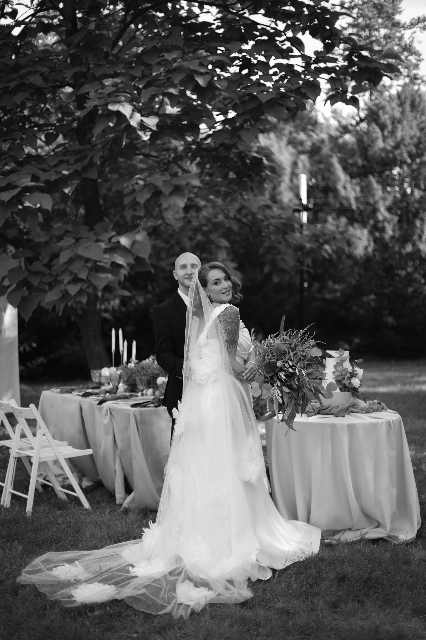 Czarno-białe zdjęcie pary młodej na tle stołu weselnego w plenerze. Panna młoda spogląda przez ramię, trzymając bukiet. Uwagę przykuwa tatuaż na jej ramieniu.