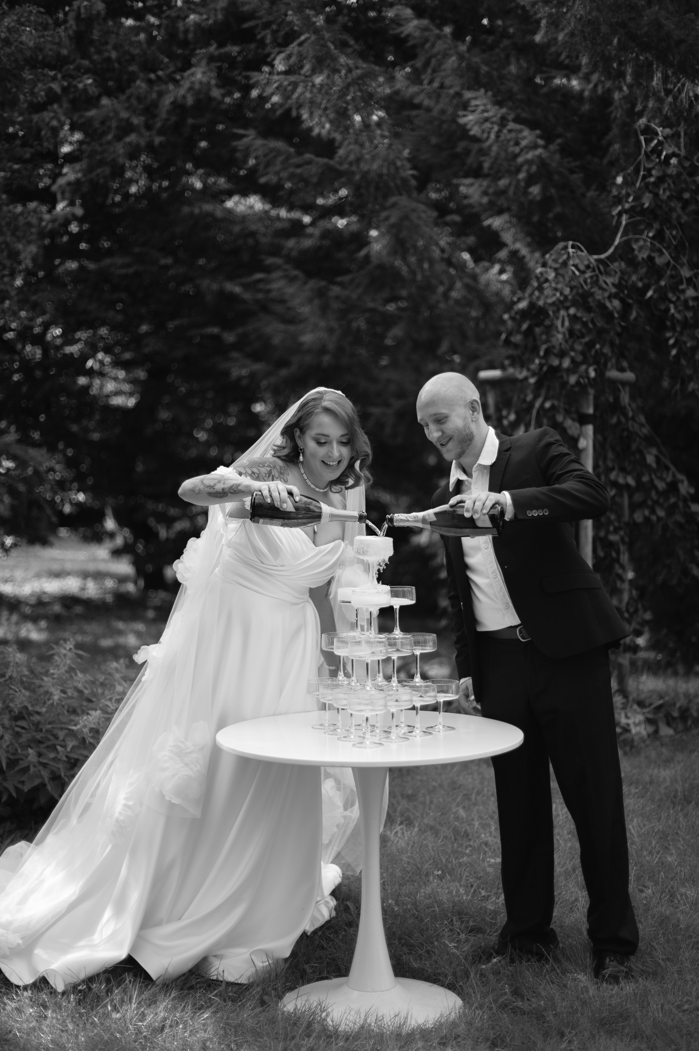 Czarno-białe zdjęcie nowożeńców w ogrodzie, nalewających szampana do piramidy z kieliszków. Panna młoda w sukni z falbanami, pan młody w garniturze. Uśmiechy na twarzach.
