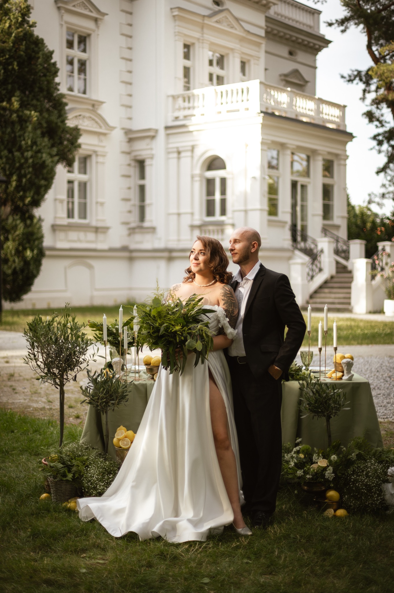 Para młoda w plenerze przed białym budynkiem. Panna młoda w sukni z rozcięciem trzyma bukiet. Dekoracje z cytryn i świec na stole.
