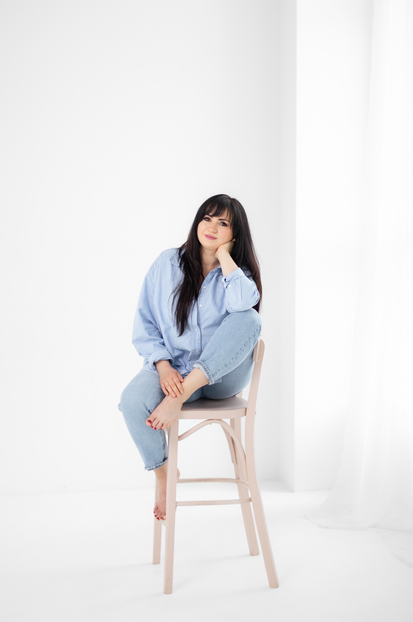 Portret kobiety w błękitnej koszuli i jeansach siedzącej na krześle na tle białego wnętrza, z nogą opartą o nogę krzesła i uśmiechem na twarzy.