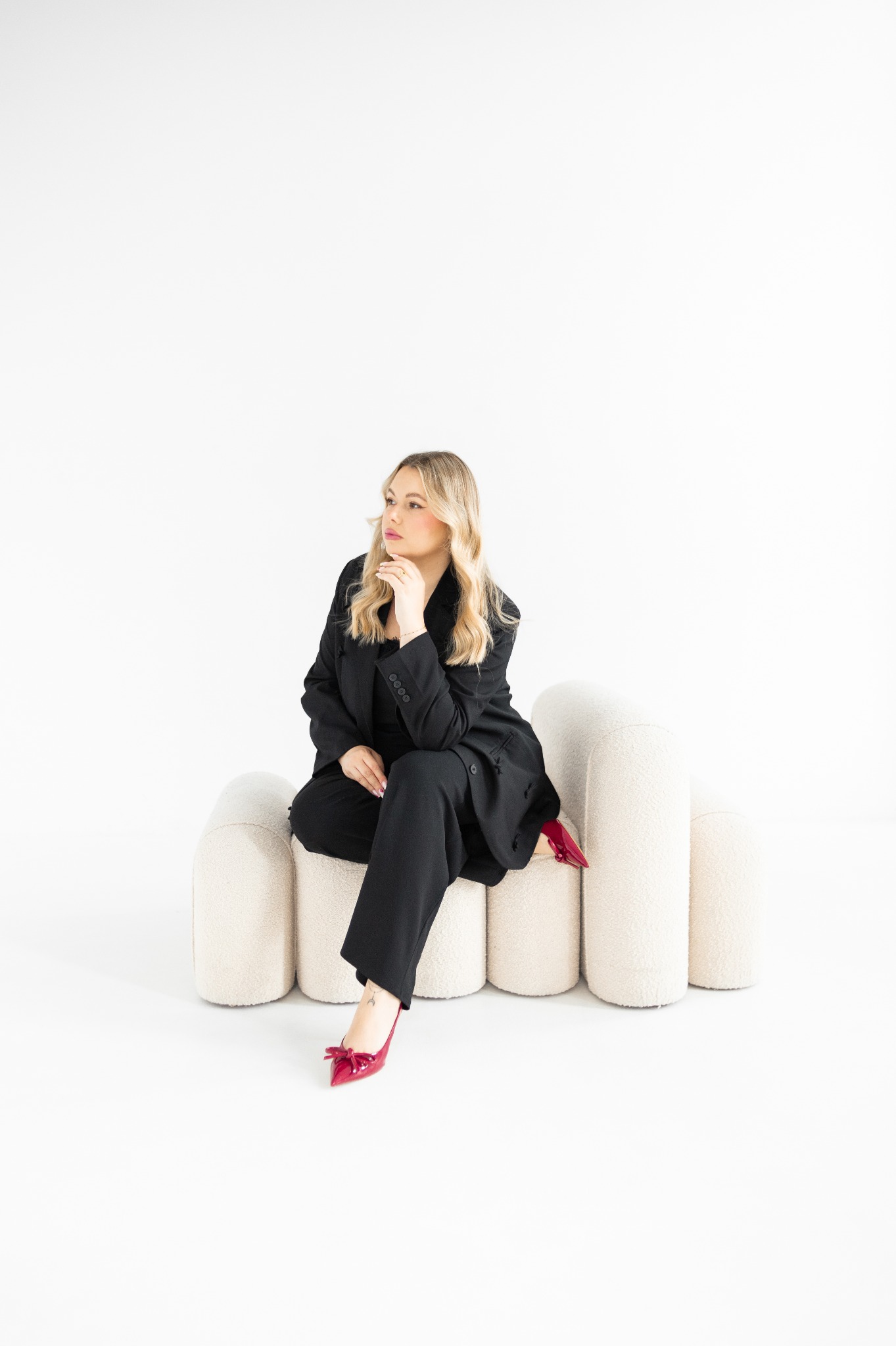 Elegancka blondynka w czarnym garniturze i czerwonych szpilkach pozuje, siedząc na designerskim fotelu na białym tle. Stonowana kolorystyka, profesjonalny look.