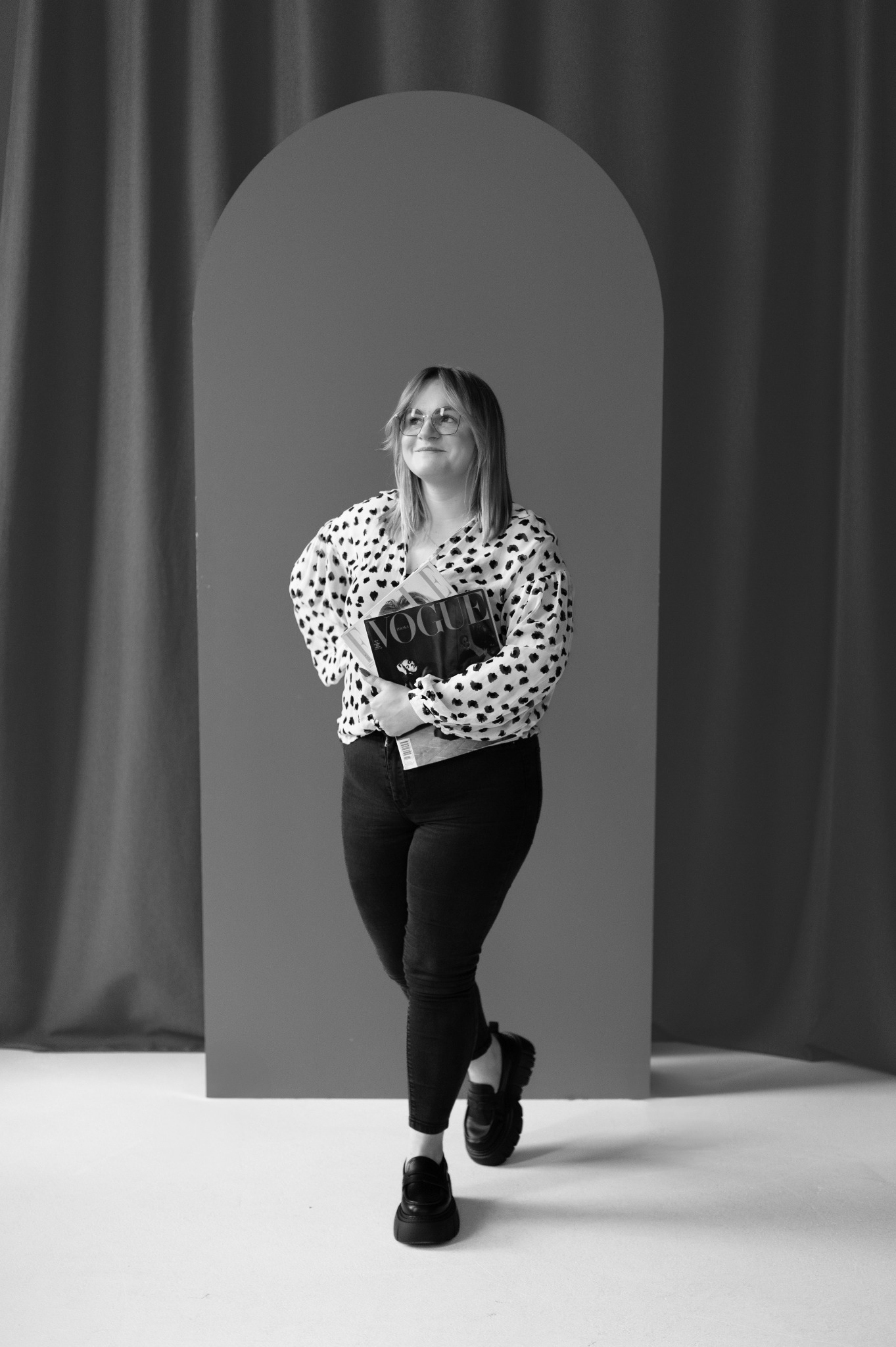 Czarno-białe zdjęcie kobiety w okularach, trzymającej magazyny 'Vogue', na tle minimalistycznej scenografii z łukiem i zasłonami. Uśmiechnięta, patrzy w górę.