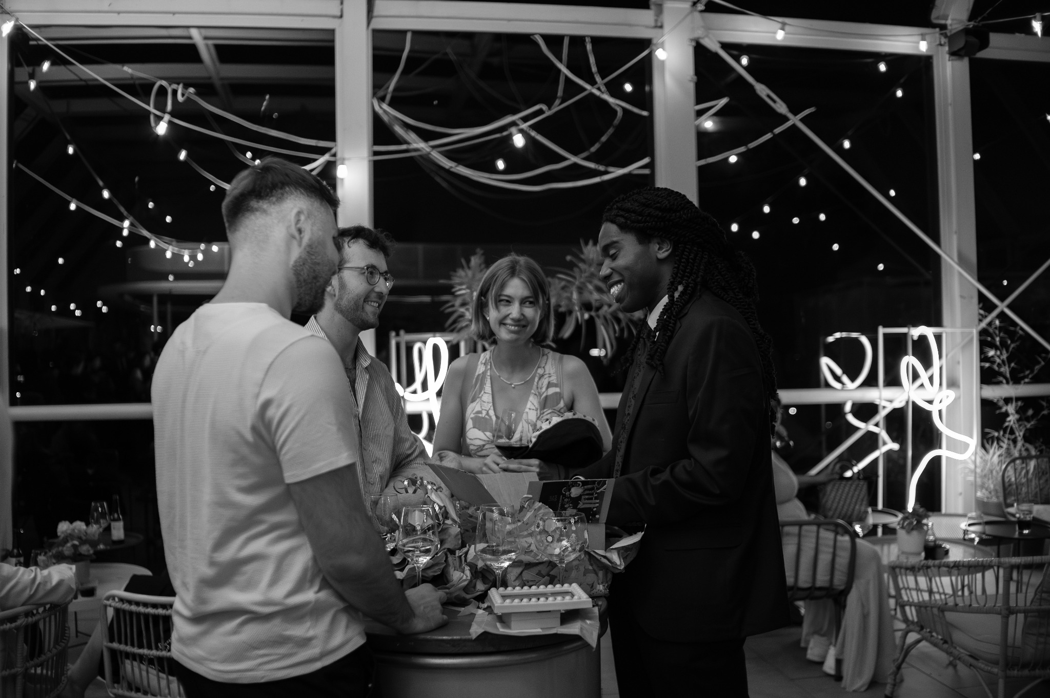 Czarno-białe zdjęcie z imprezy: grupa ludzi rozmawia przy barze, w tle dekoracje świetlne i okna. Uśmiechnięta kobieta trzyma kieliszek wina.