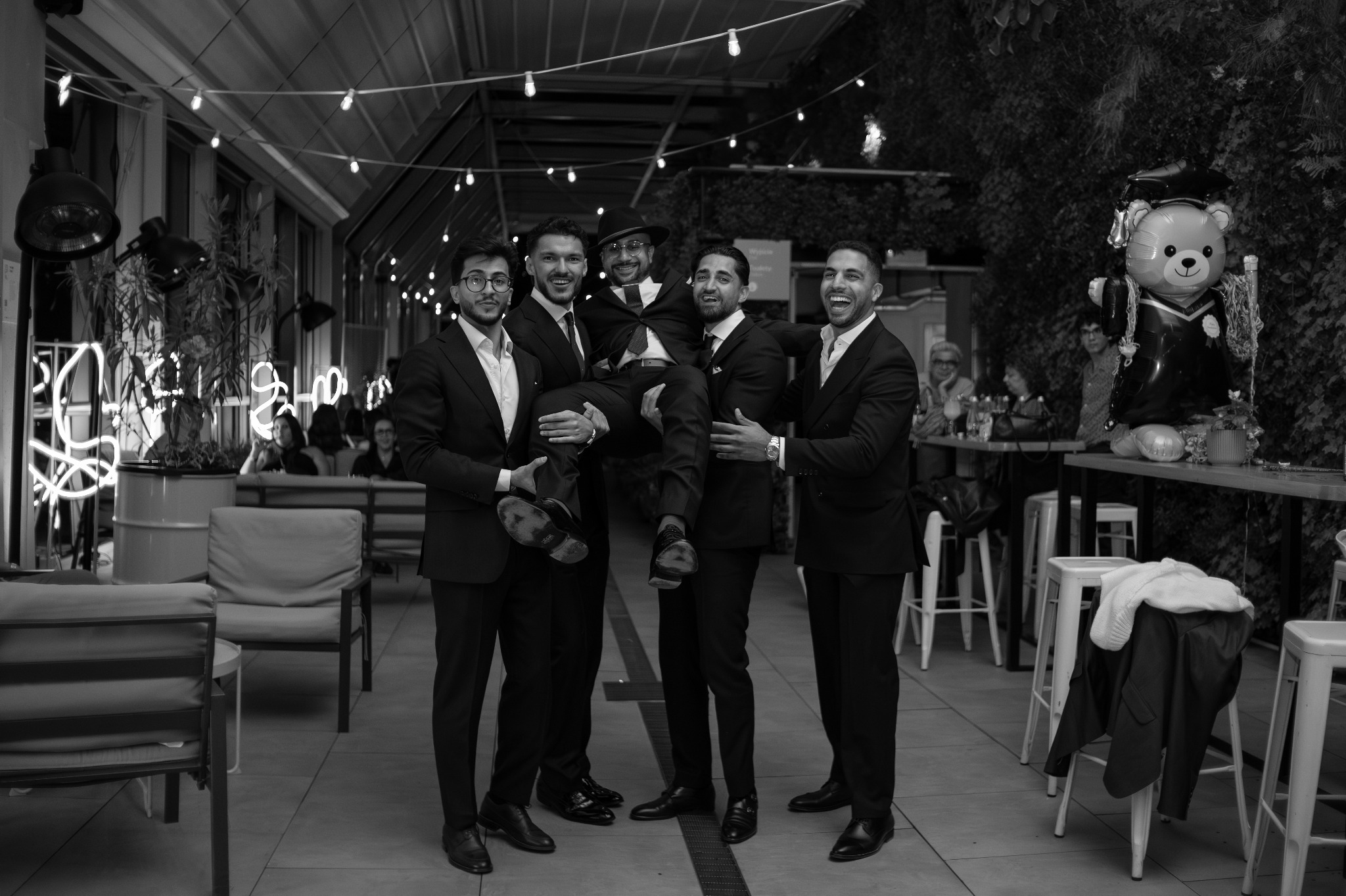 Czarno-białe zdjęcie grupy pięciu mężczyzn w garniturach, czterech z nich trzymającego jednego na rękach. W tle dekoracje i goście.