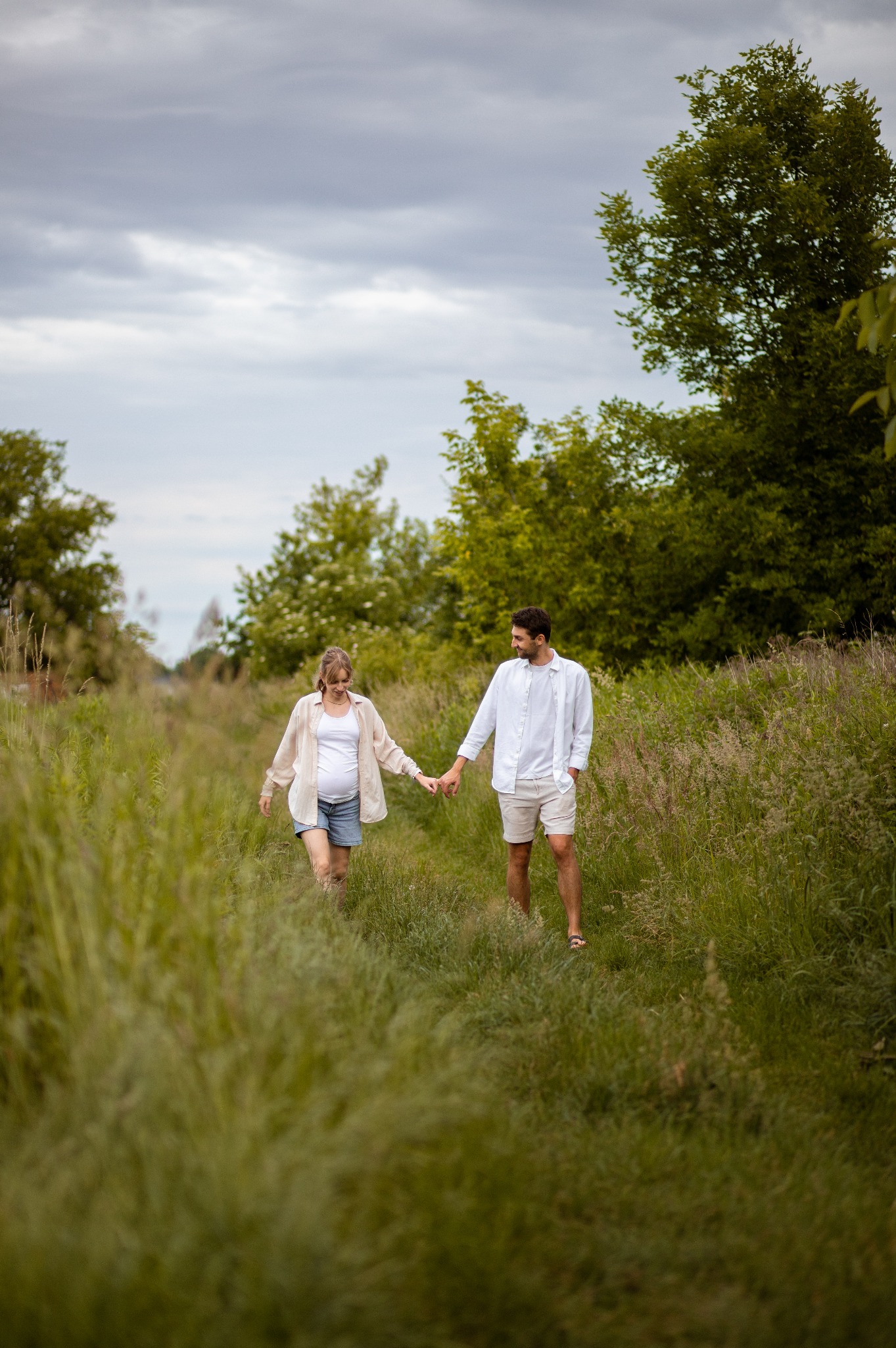 Sesja ciążowa we Wrocławiu: Przyszła mama trzyma za rękę partnera, spacerując pośród wysokich traw. Romantyczna sceneria i naturalne światło.
