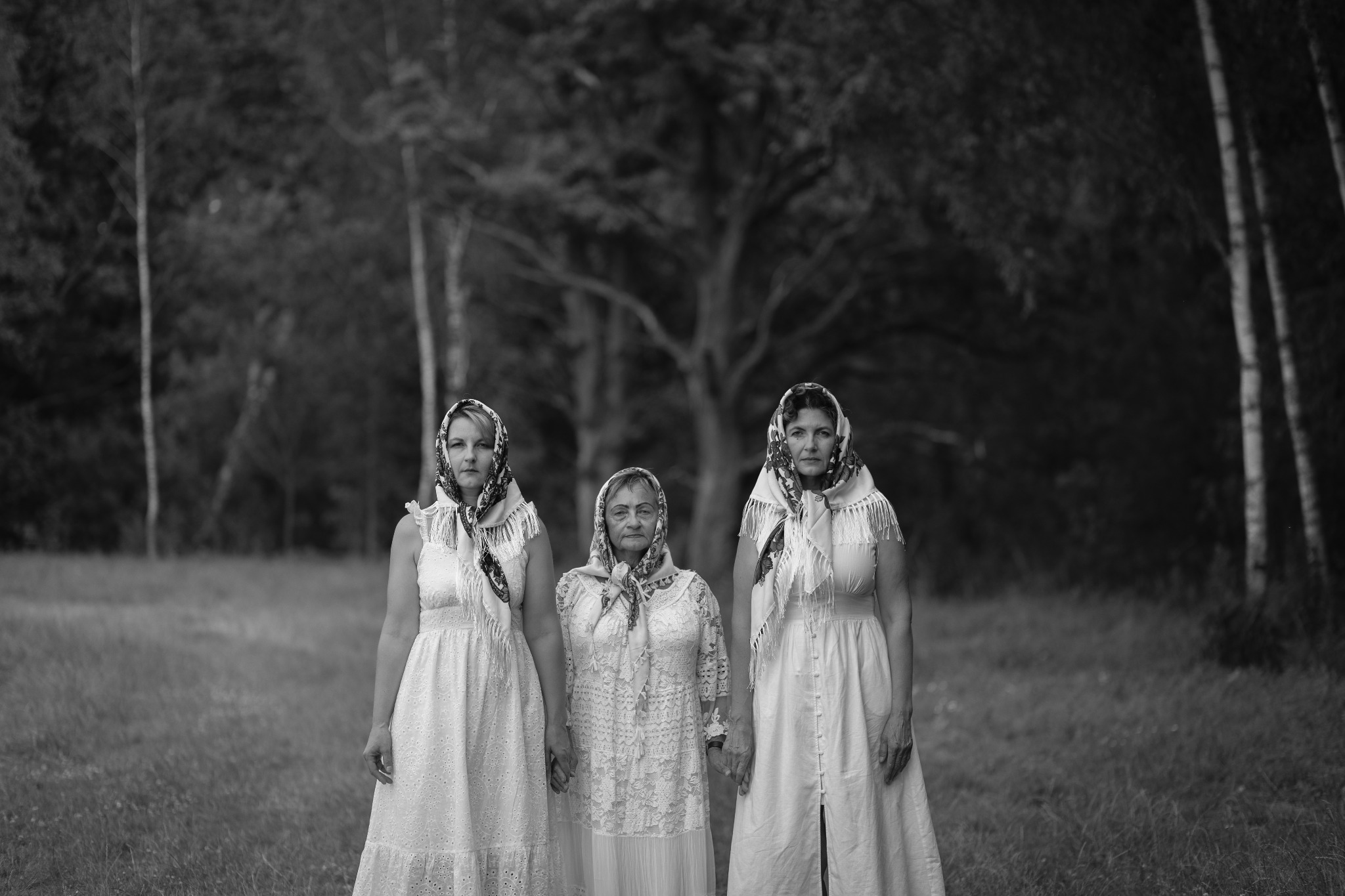 Czarno-białe zdjęcie trzech kobiet w białych sukienkach i chustach na głowach, stojących na tle drzew w plenerze. Ujęcie centralne, statyczne.