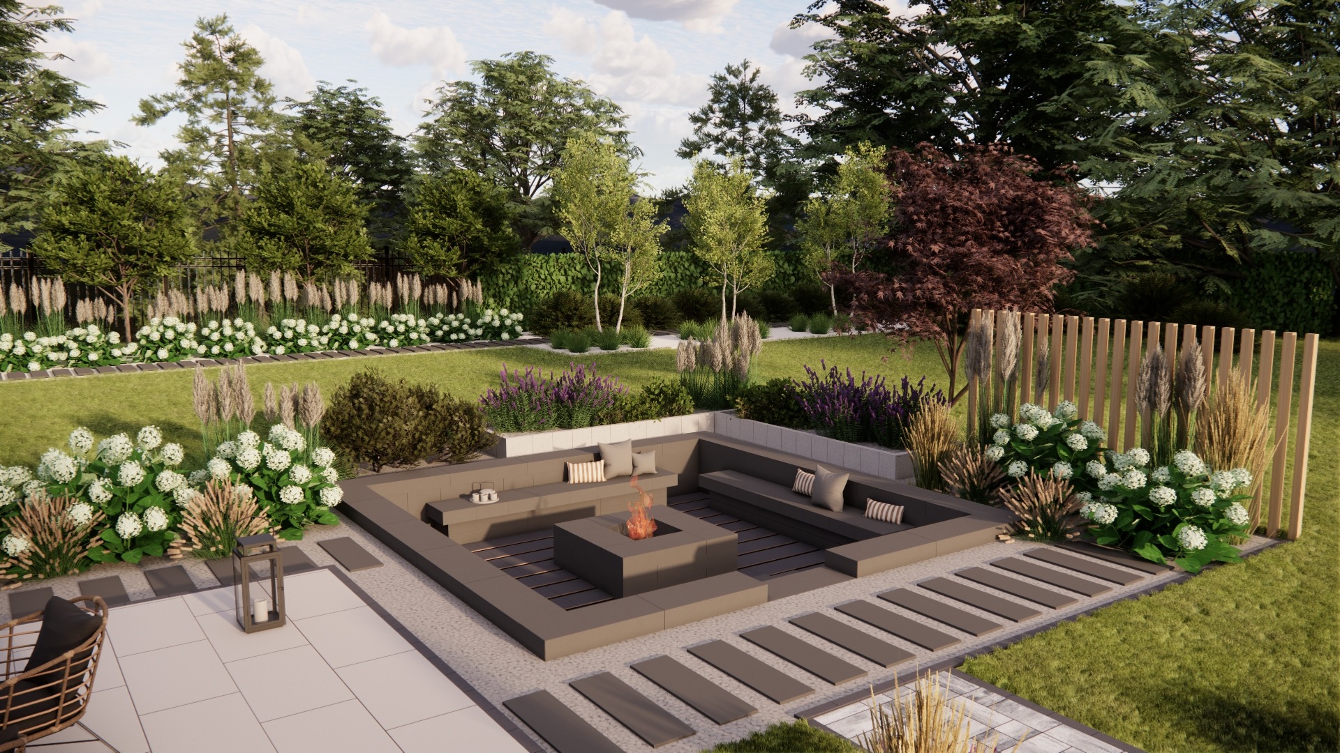 Ogród z paleniskiem otoczonym siedziskami, niską drewnianą palisadą i rabatami z kwitnącymi krzewami oraz trawami ozdobnymi, widok z perspektywy tarasu.