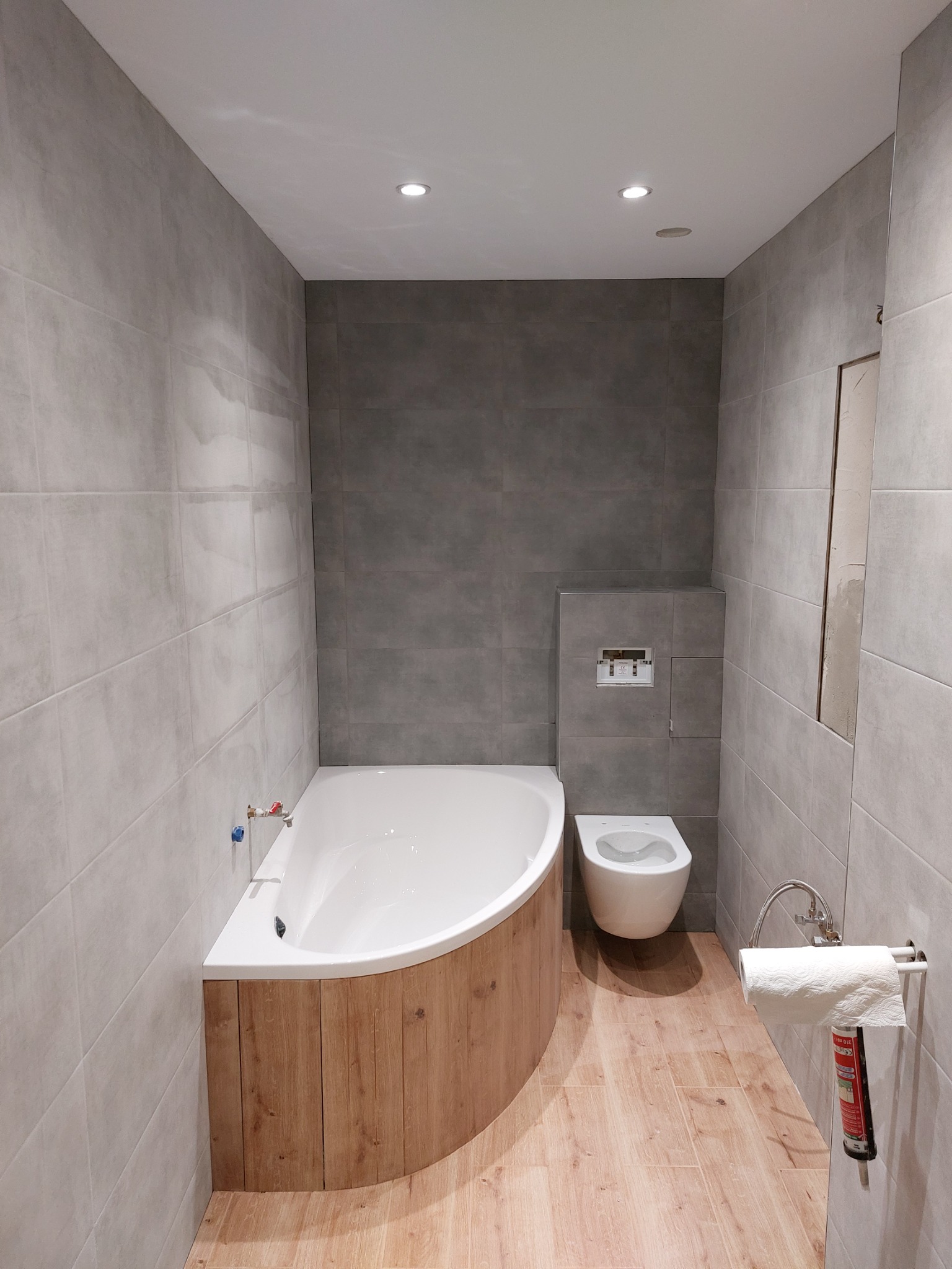 Nowoczesna łazienka z szarymi płytkami, drewnopodobną obudową wanny narożnej i podwieszaną toaletą, uzupełniona o drewnopodobne płytki podłogowe i minimalistyczny uchwyt na papier toaletowy...