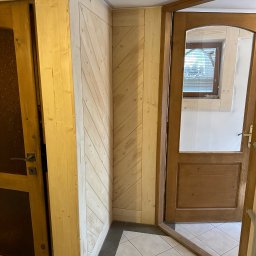 Ufarbiorza - Wnętrze z drewnianą boazerią ułożoną pod kątem, drewniane drzwi z szybą w stylu rustykalnym, widoczny fragment podłogi wyłożonej płytkami.