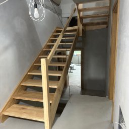 Ufarbiorza - Surowe, jasne drewniane schody w trakcie montażu w nowym domu, widoczne kable i surowy beton, prowizoryczna żarówka.