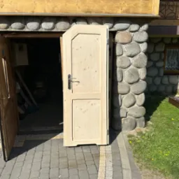 Otwarta, surowa drewniana dwuskrzydłowa brama z czarną klamką, osadzona w kamiennej ścianie budynku z drewnianym wykończeniem, na tle brukowanej ścieżki i trawy.