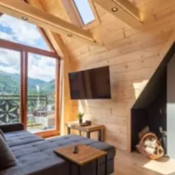 Drewniane wnętrze w stylu góralskim z dużym oknem balkonowym i widokiem na góry. Drewniane ściany, sufit ze świetlikami i nowoczesny telewizor.