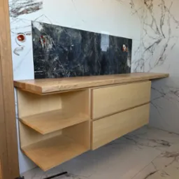 Drewniana szafka łazienkowa z blatem z jasnego drewna, z otwartymi półkami i szufladami, na tle marmurowych płytek. Widoczne narzędzia stolarskie na podłodze.