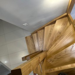 Ufarbiorza - Drewniane schody spiralne z jasnego drewna dębowego, widoczne z góry, z fragmentem białej podłogi i sufitu. U dołu kadru fragment buta.