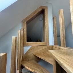 Ufarbiorza - Drewniane schody z balustradą i nietypowym oknem w kształcie trapezu wkomponowanym w ścianę poddasza, widok z dołu, naturalne oświetlenie.