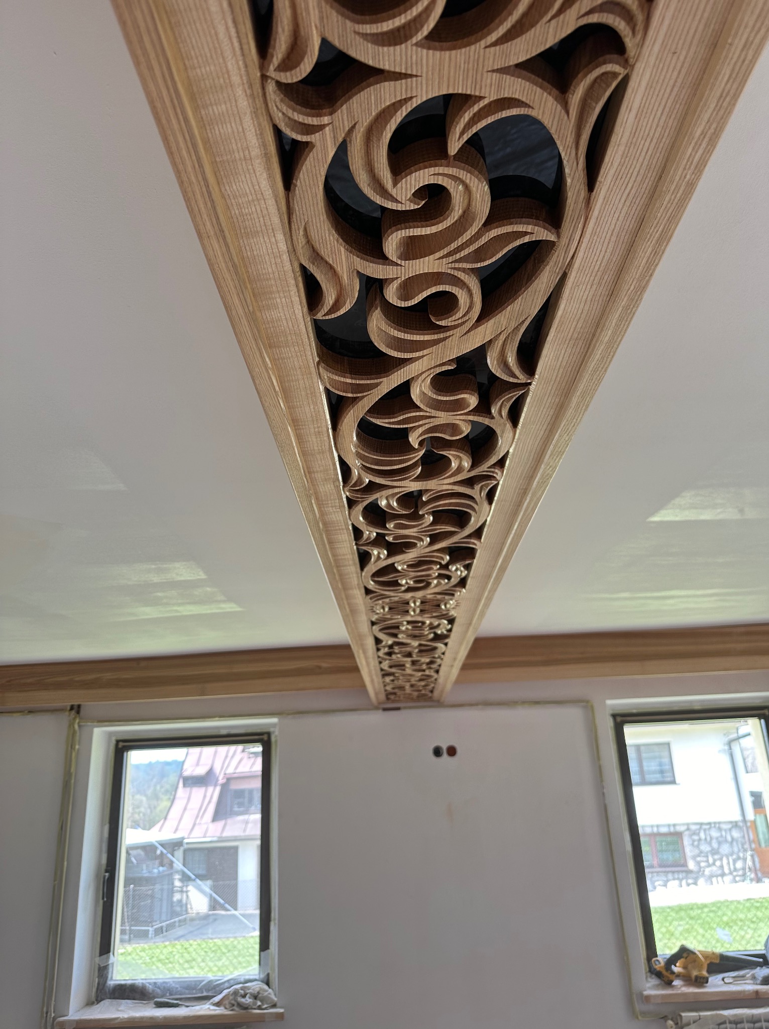 Dekoracyjny, ażurowy element z drewna zdobiący sufit w jasnym pomieszczeniu, widoczne okna z widokiem na zewnątrz.