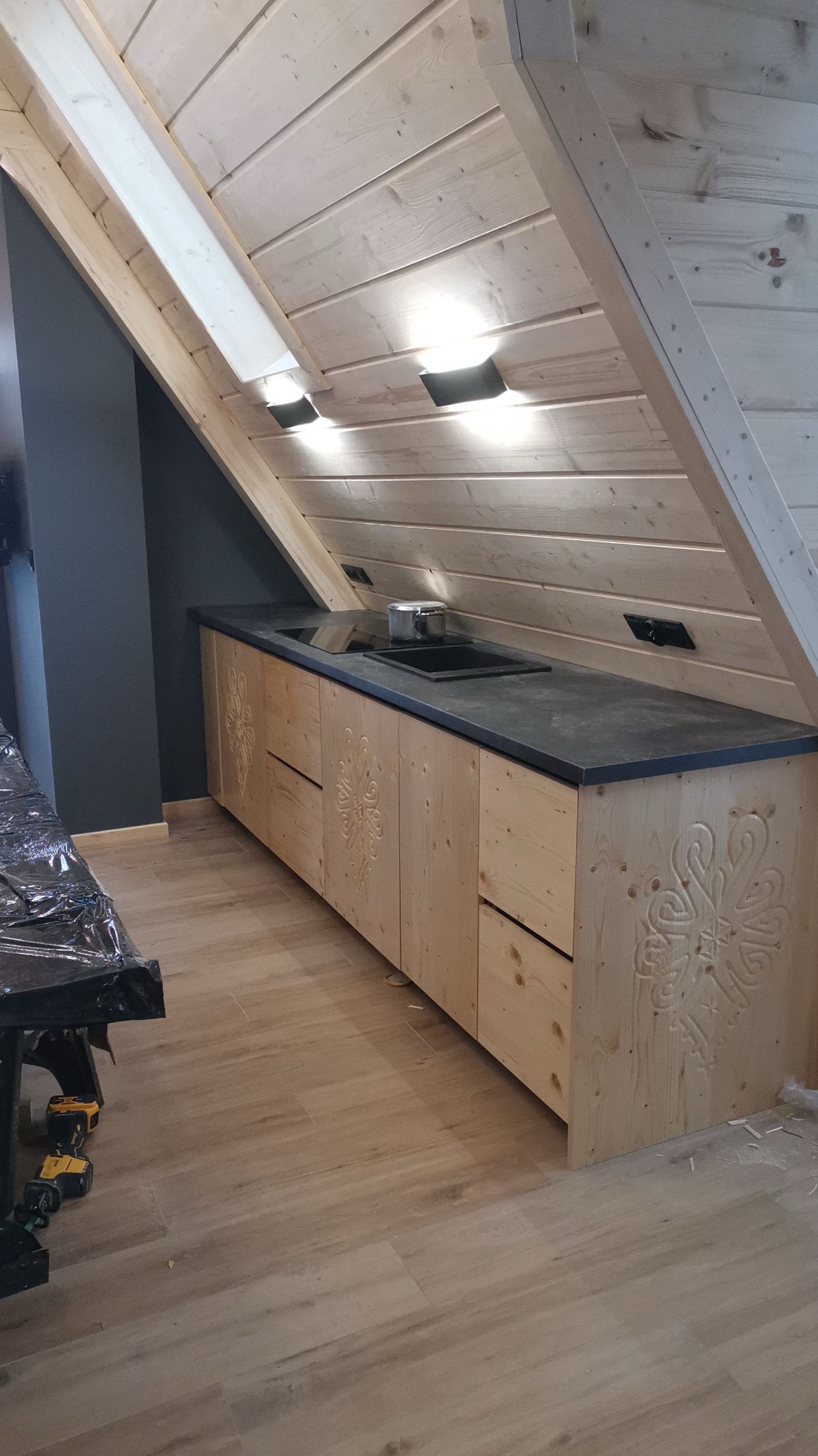 Kuchenna zabudowa z jasnego drewna z góralskimi wzorami pod skośnym dachem z drewnianą boazerią, czarny blat z wbudowaną płytą indukcyjną i zlewozmywakiem, fragment stołu roboczego z narzędziami...