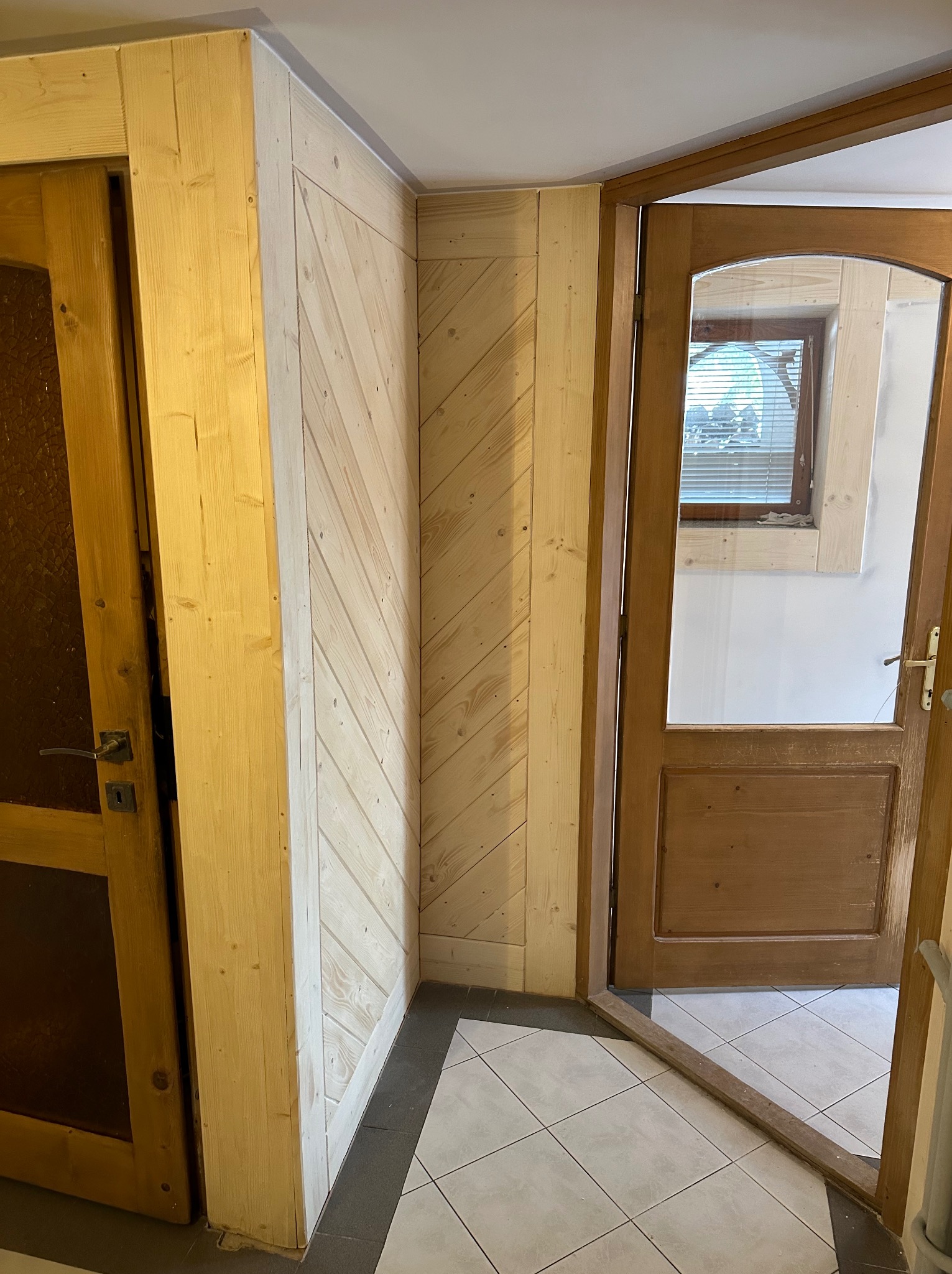 Wnętrze z drewnianą boazerią ułożoną pod kątem, drewniane drzwi z szybą w stylu rustykalnym, widoczny fragment podłogi wyłożonej płytkami.