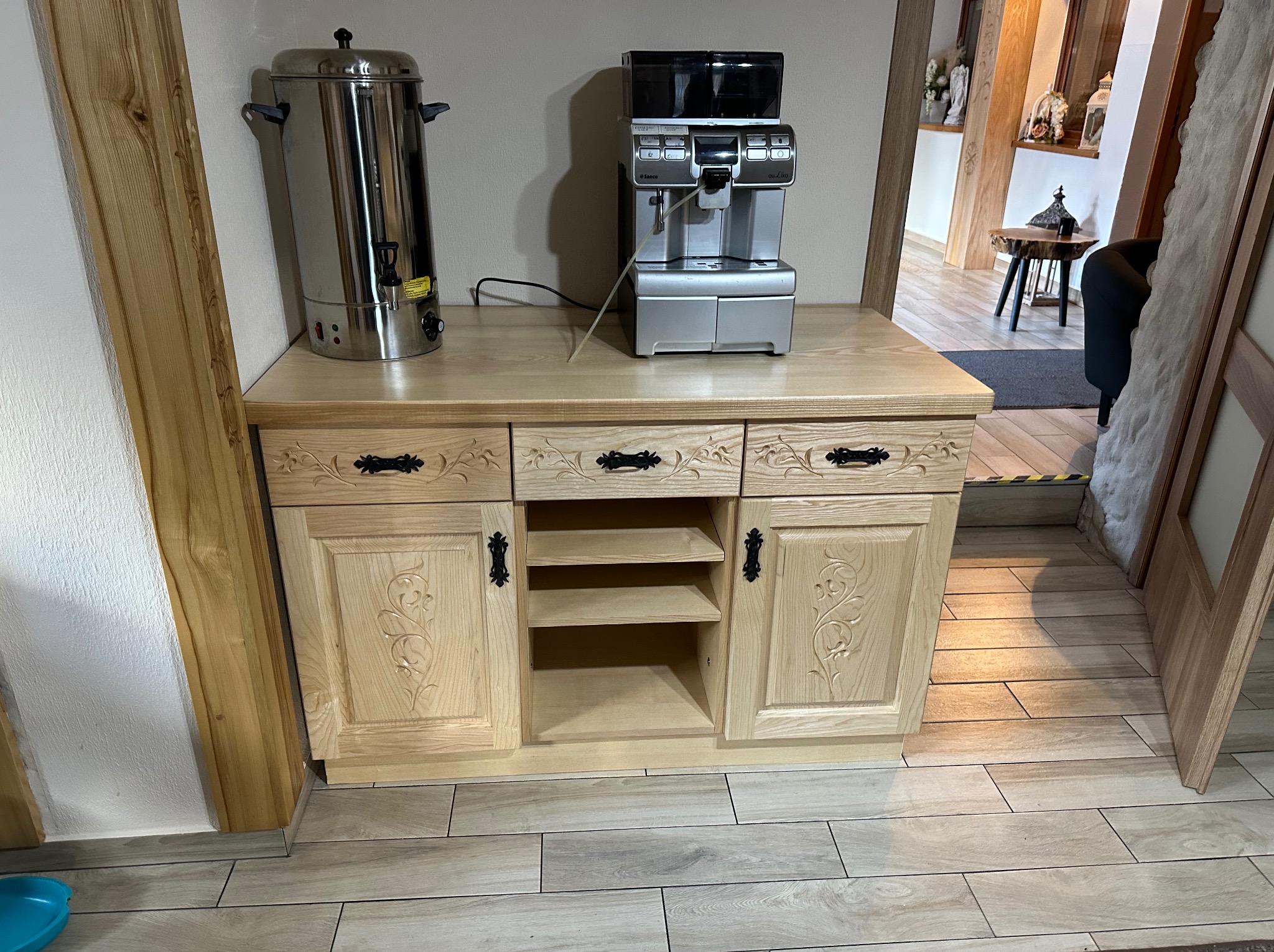 Jasna, drewniana szafka z rzeźbionymi frontami, na której stoją ekspres do kawy i termos, widok na wnętrze domu w tle.