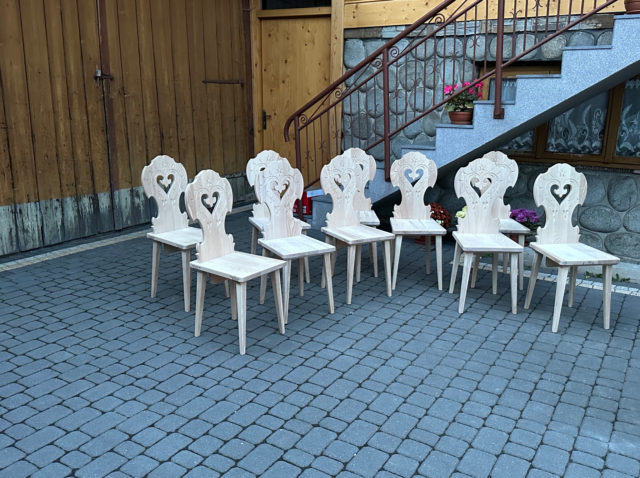 Siedem surowych, drewnianych krzeseł z wyciętym motywem serca w oparciu, ustawionych na brukowanym dziedzińcu przed drewnianymi drzwiami i kamiennymi schodami z kwiatami.