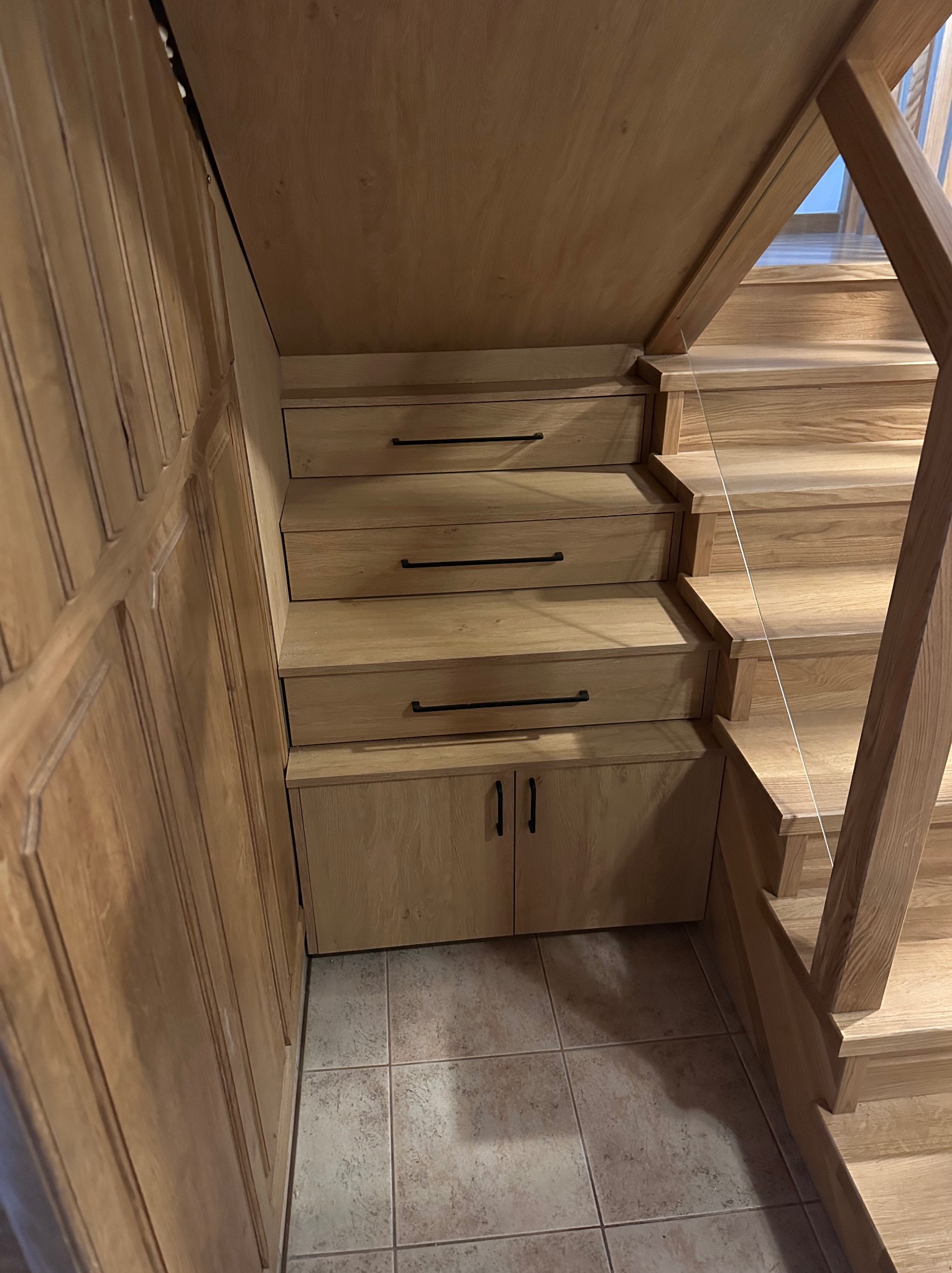 Drewniane schody z szufladami i szafką pod spodem, jasne drewno, czarne uchwyty, płytki podłogowe.