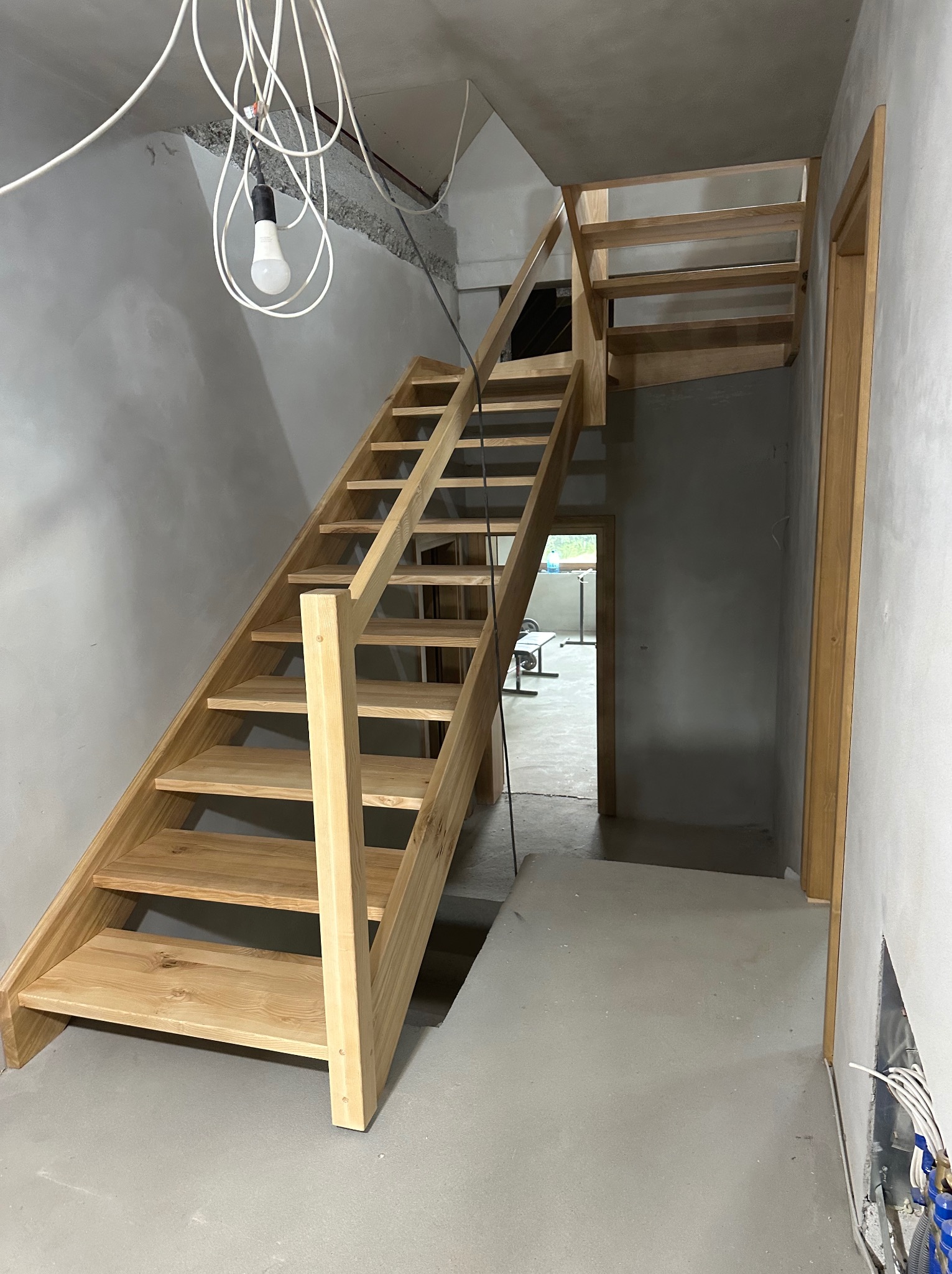 Surowe, jasne drewniane schody w trakcie montażu w nowym domu, widoczne kable i surowy beton, prowizoryczna żarówka.